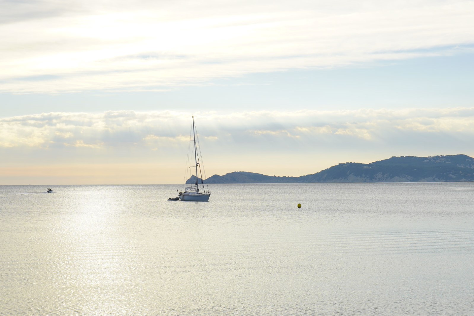 Vista del mar Mediterráneo cerca del puerto de Jávea, con un velero navegando bajo una luz suave y la silueta de la costa al fondo.