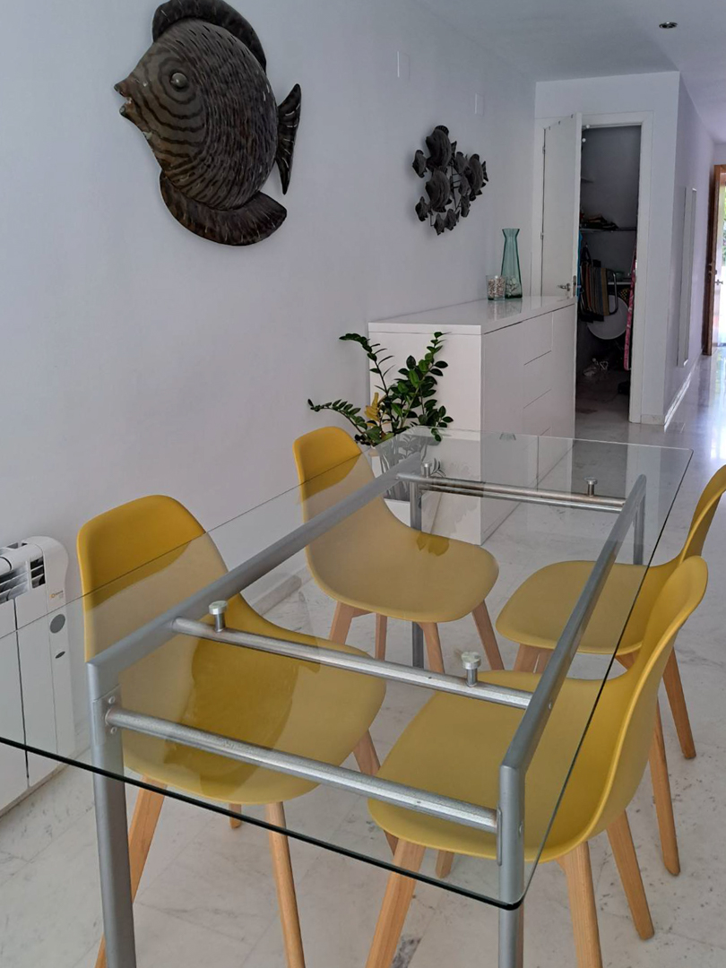 Zona de comedor con mesa de cristal y sillas amarillas sobre suelo de mármol. La estancia incluye un aparador blanco, radiador eléctrico y decoración mural de inspiración marina.