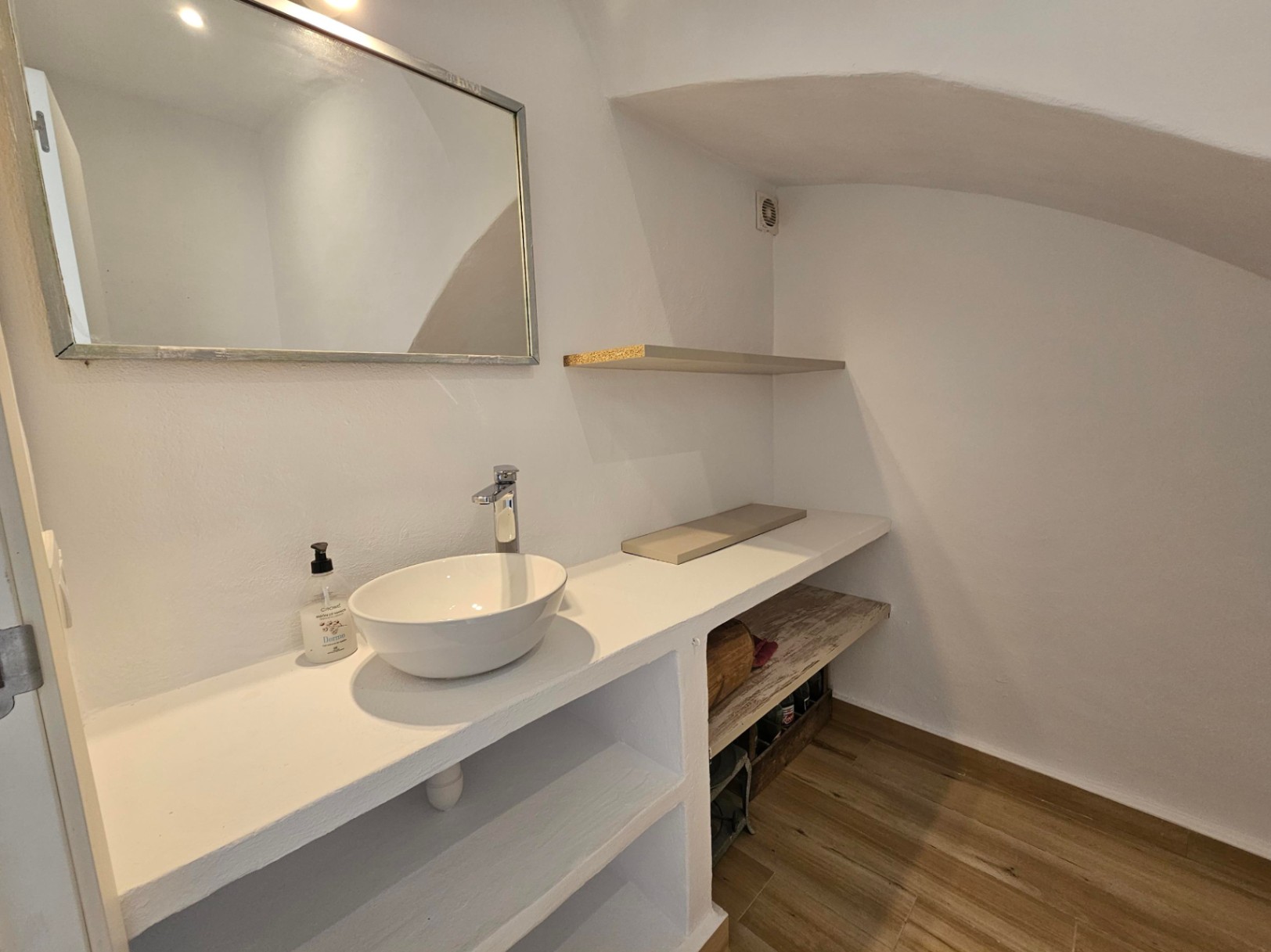 Aseo de cortesía en planta baja con lavabo sobre encimera blanca, estanterías integradas y techo inclinado. Suelo de gres efecto madera y espejo rectangular.