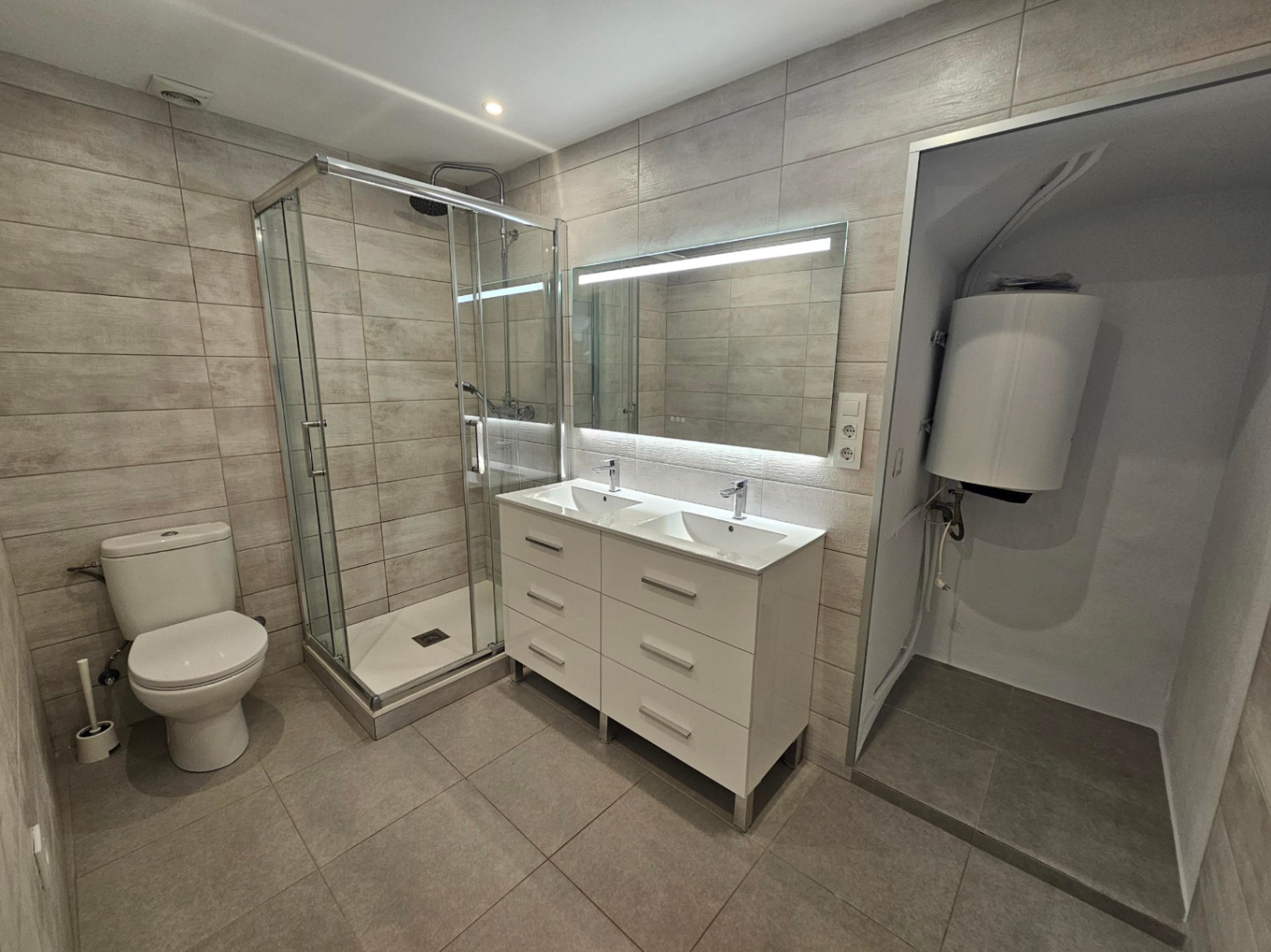 Baño moderno con mueble de doble lavabo, espejo LED y ducha con mampara de cristal. Presenta azulejos cerámicos en tonos neutros y zona técnica para el termo.