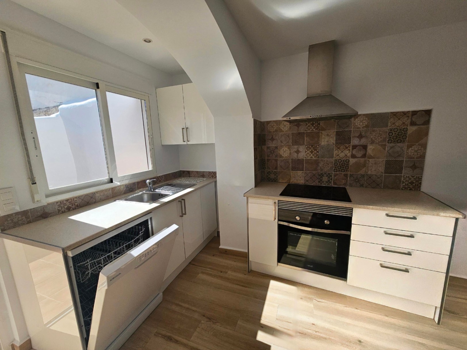 Cocina moderna reformada con muebles blancos, encimera clara y frontal de azulejos hidráulicos. Incluye electrodomésticos integrados y suelos de tarima de madera.