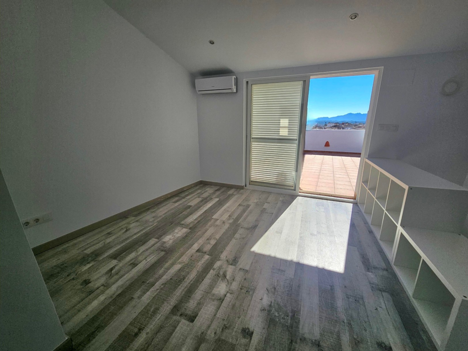 Despacho luminoso con suelo laminado, aire acondicionado y acceso directo a una terraza privada con vistas a la montaña. Incluye estantería blanca y focos empotrados.