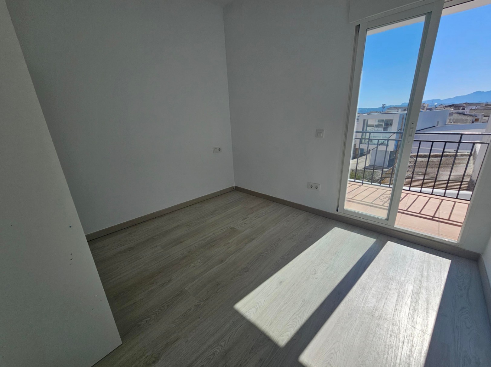 Dormitorio luminoso con suelos laminados de madera clara, paredes blancas y acceso directo a un balcón privado con vistas despejadas al entorno urbano y las montañas.