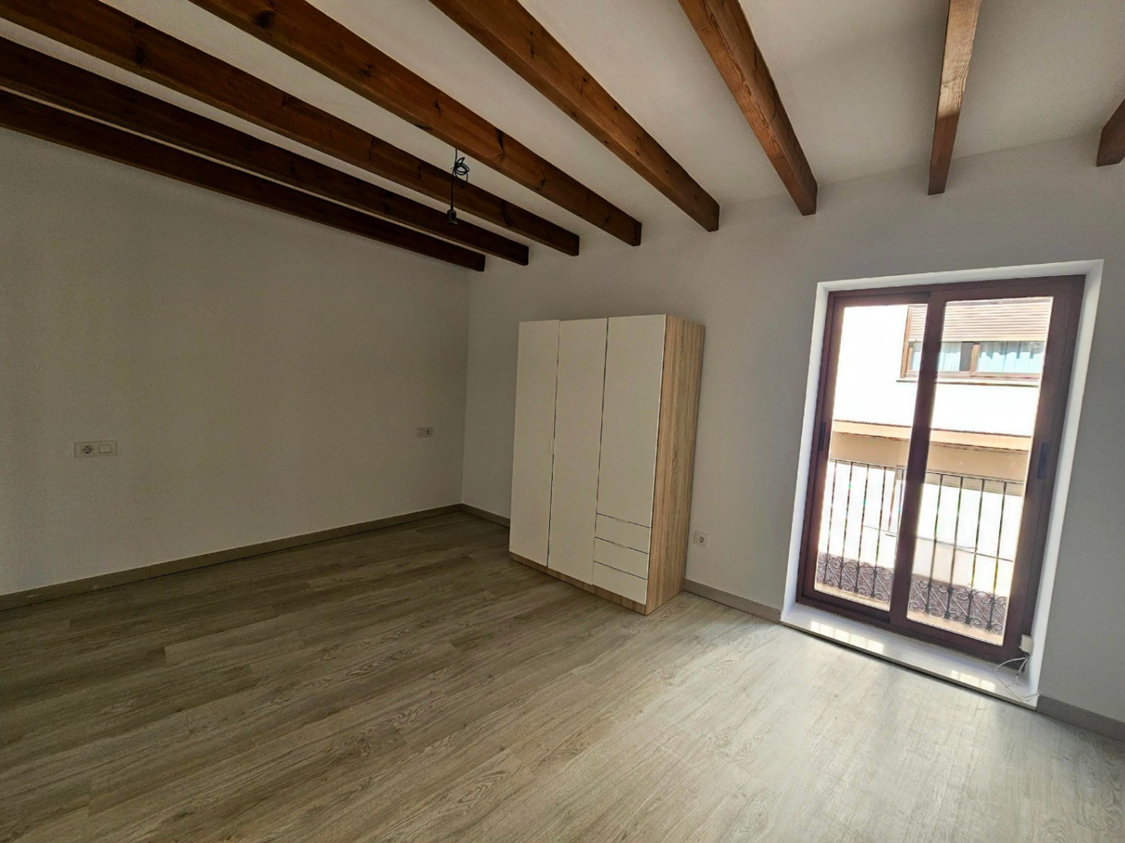 Dormitorio luminoso con vigas de madera vista, suelo laminado claro y acceso directo a un balcón privado a través de puertas de doble acristalamiento.