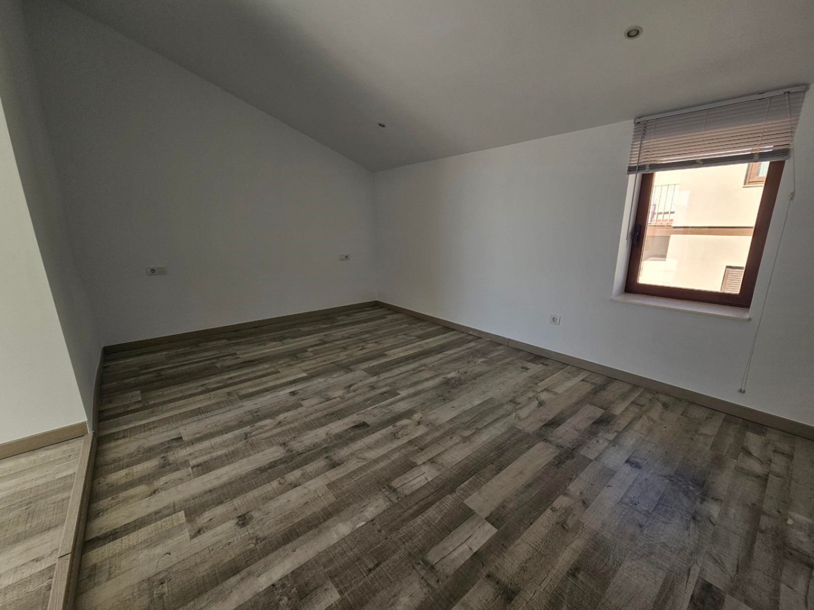 Habitación diáfana con suelos de tarima de efecto madera rústica, paredes blancas y techo inclinado con iluminación empotrada. Incluye ventana con persiana.