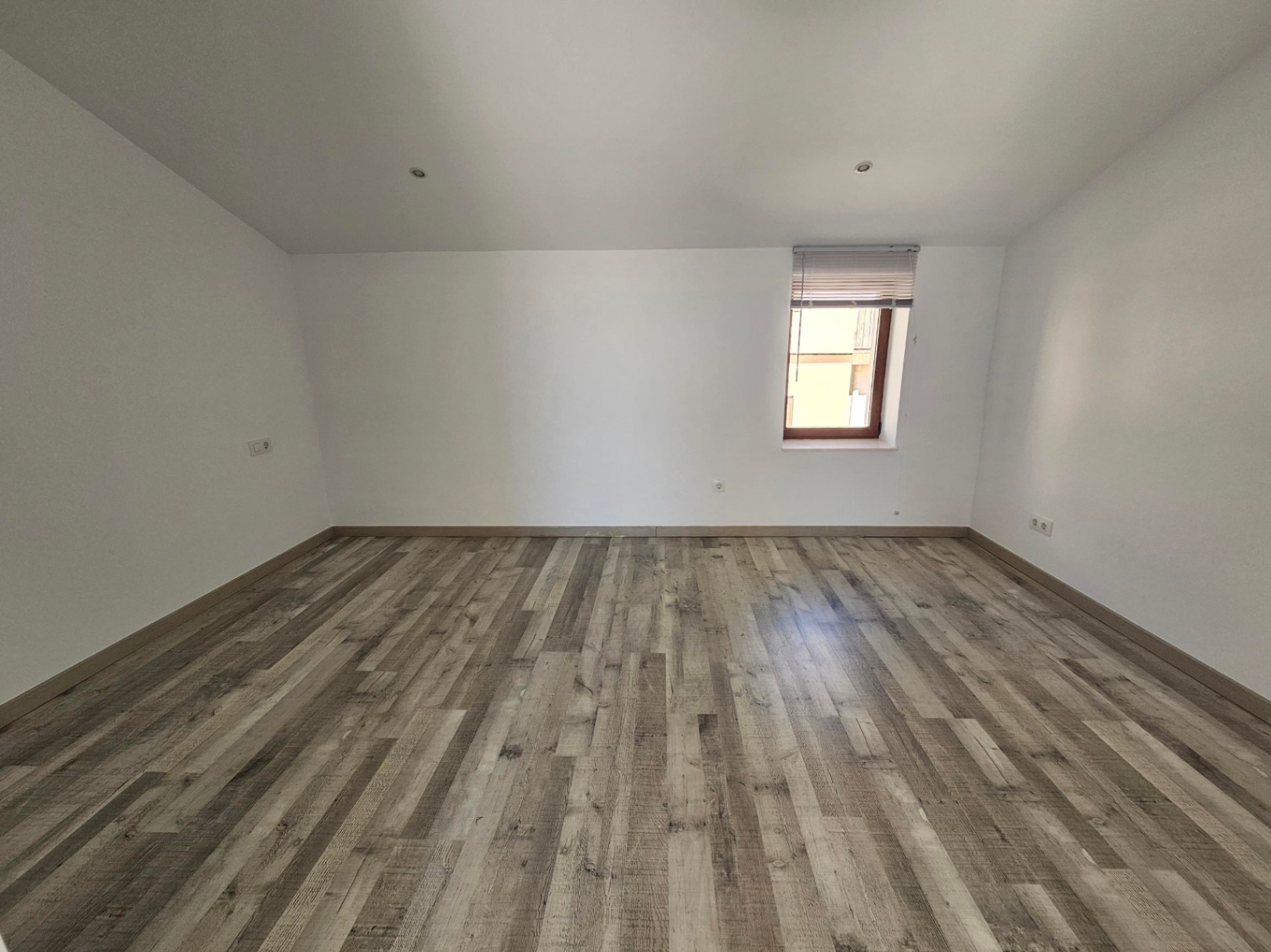 Habitación diáfana con techos abuhardillados, suelos de tarima laminada en tonos grises y paredes blancas. Incluye focos empotrados y una ventana con marco de madera.