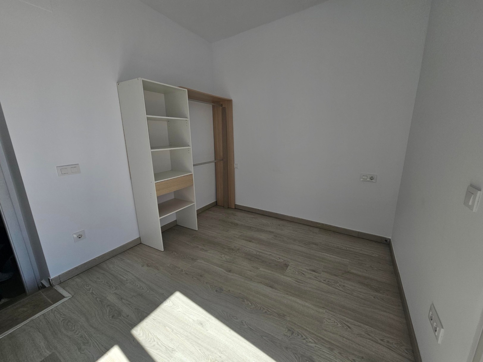 Habitación luminosa con suelos de tarima clara, paredes blancas y un práctico armario abierto con estanterías y zona de colgar.