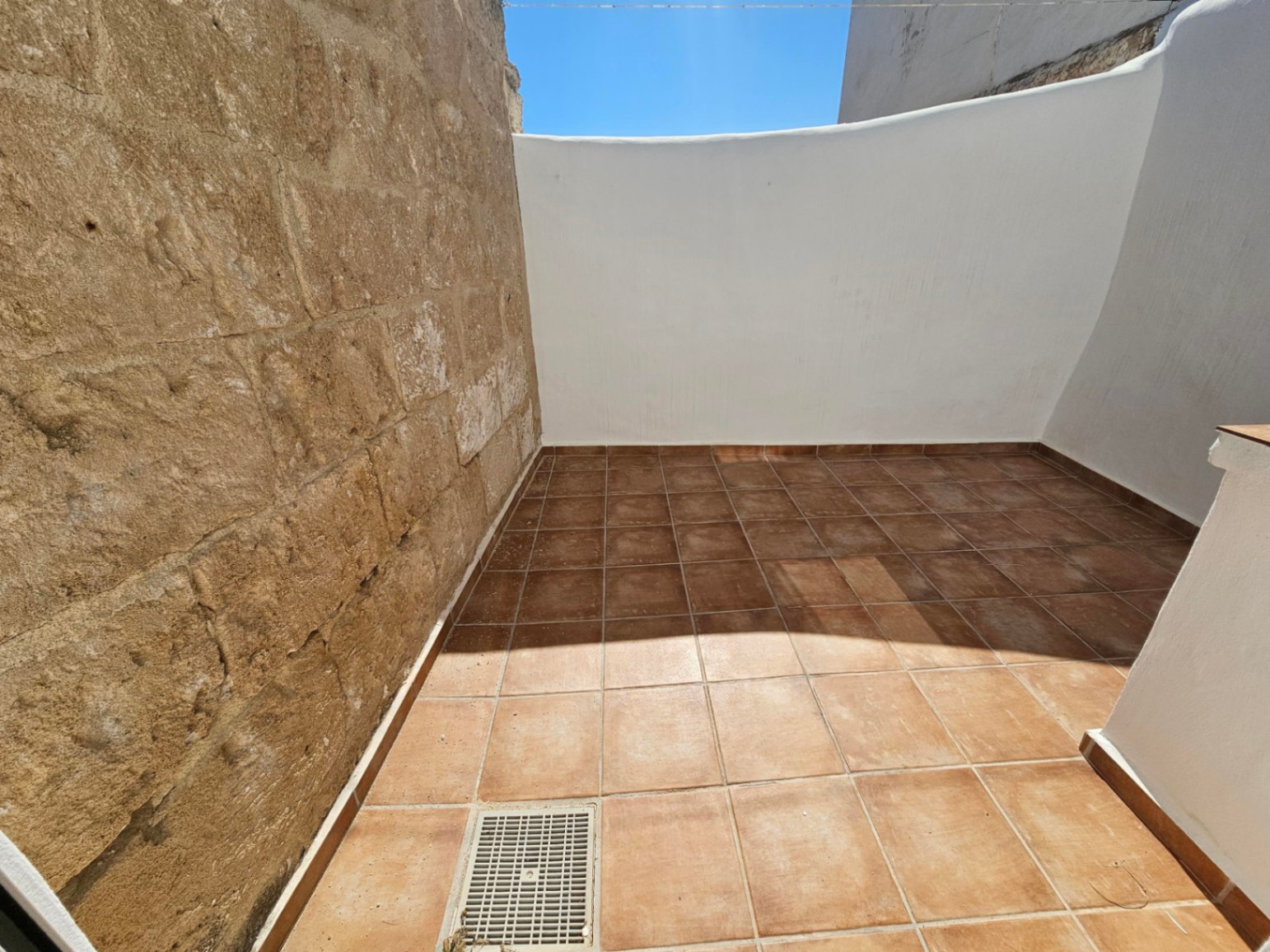 Patio exterior privado con suelo de baldosas cerámicas terracota, muro de piedra natural original y paredes blancas bajo el cielo despejado.
