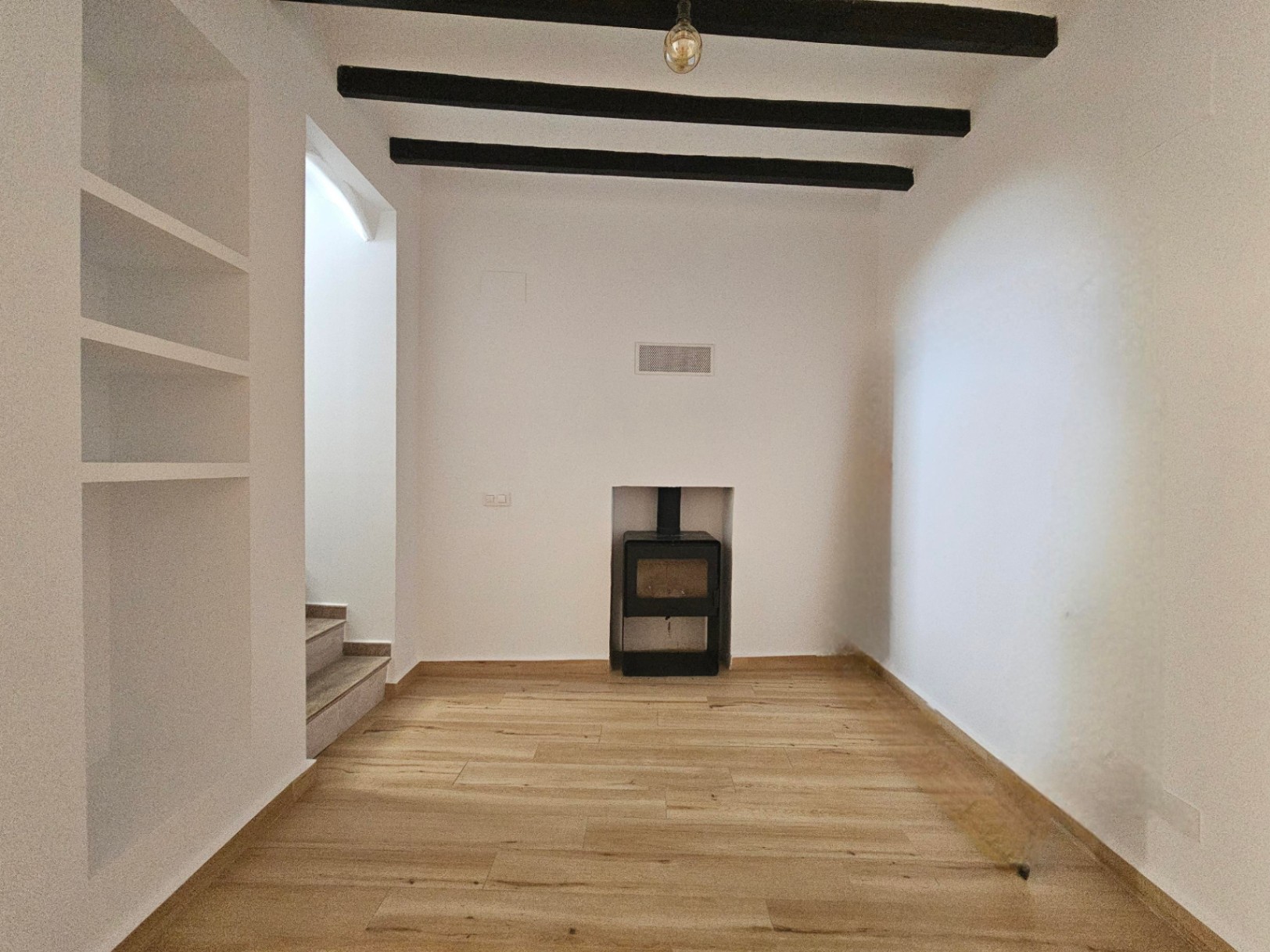 Salón con quemador de leña encastrado, vigas de madera oscura en el techo y suelos de tarima clara. Incluye estantería de obra blanca y acceso lateral a la escalera.