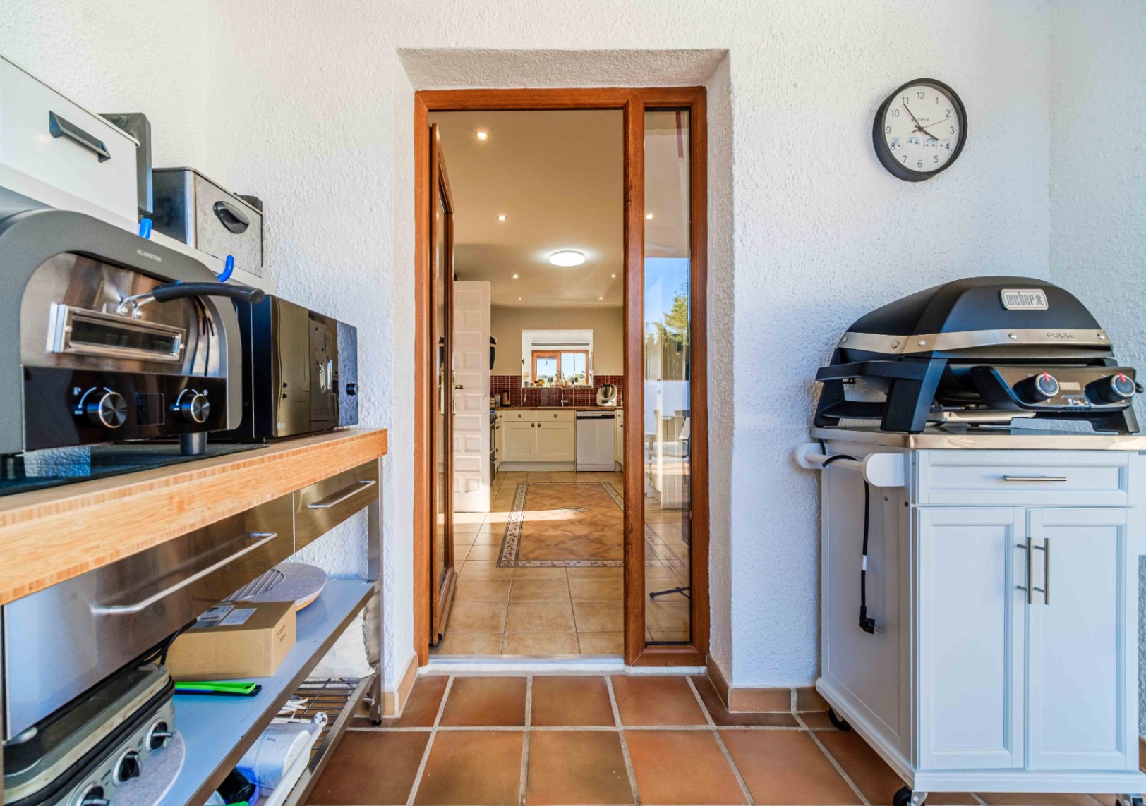 Área de cocina exterior equipada con barbacoa eléctrica Weber y estanterías de acero, con acceso directo a la cocina interior a través de una puerta acristalada.