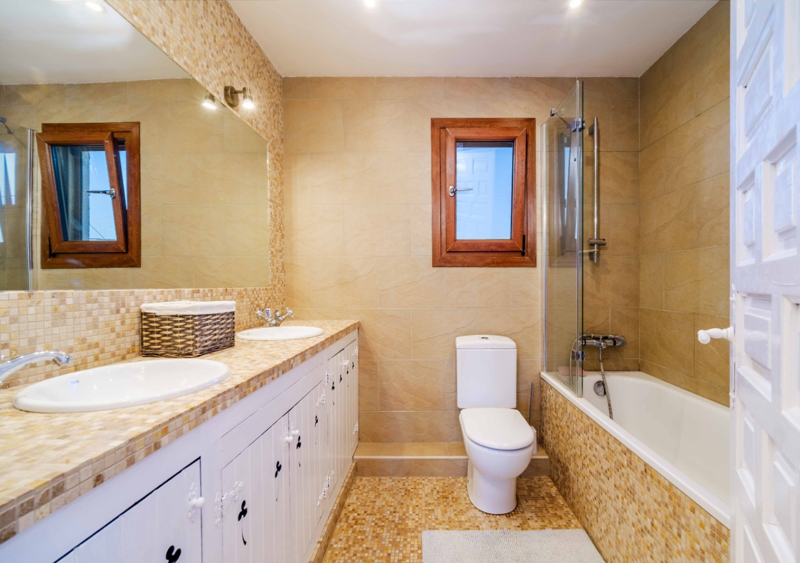 Baño completo con lavabo doble, encimera de mosaico y muebles blancos. Incluye bañera con mampara de cristal, azulejos en tonos crema y ventana con marco de madera.