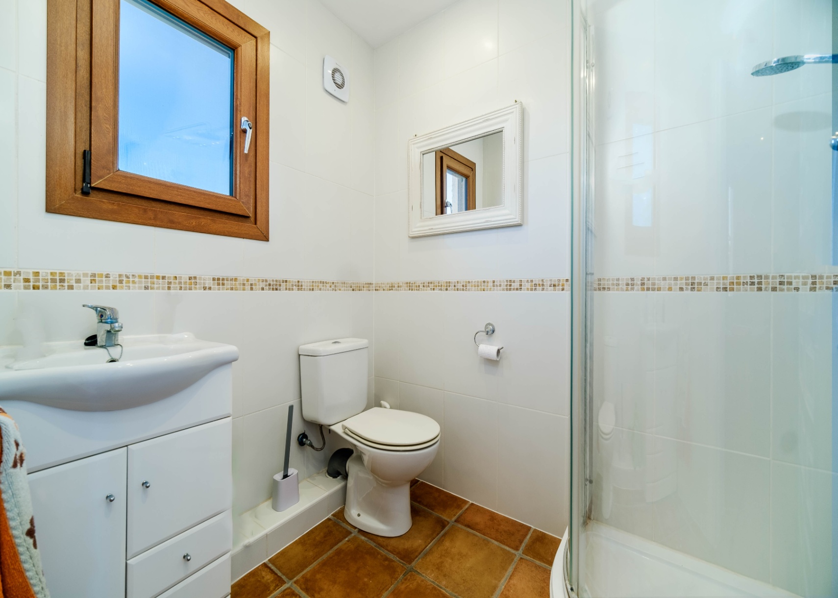 Baño con paredes alicatadas en blanco, cenefa de mosaico y suelo de terracota. Incluye ducha con mampara de cristal, mueble lavabo y una ventana con marco de madera.