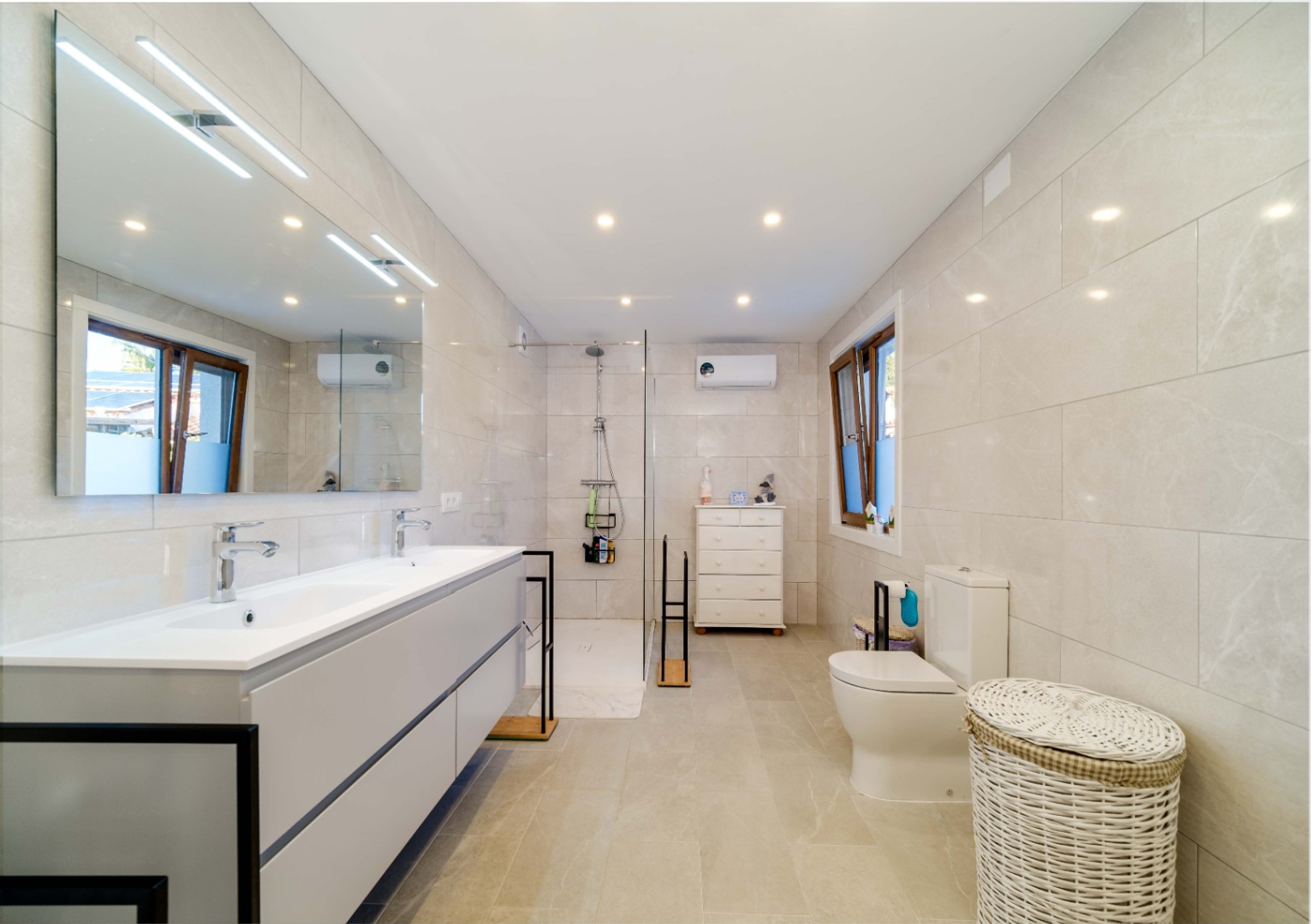 Baño moderno con lavabo doble, mueble suspendido gris, ducha a ras de suelo con mampara de cristal y paredes revestidas en gres porcelánico de tonos neutros.