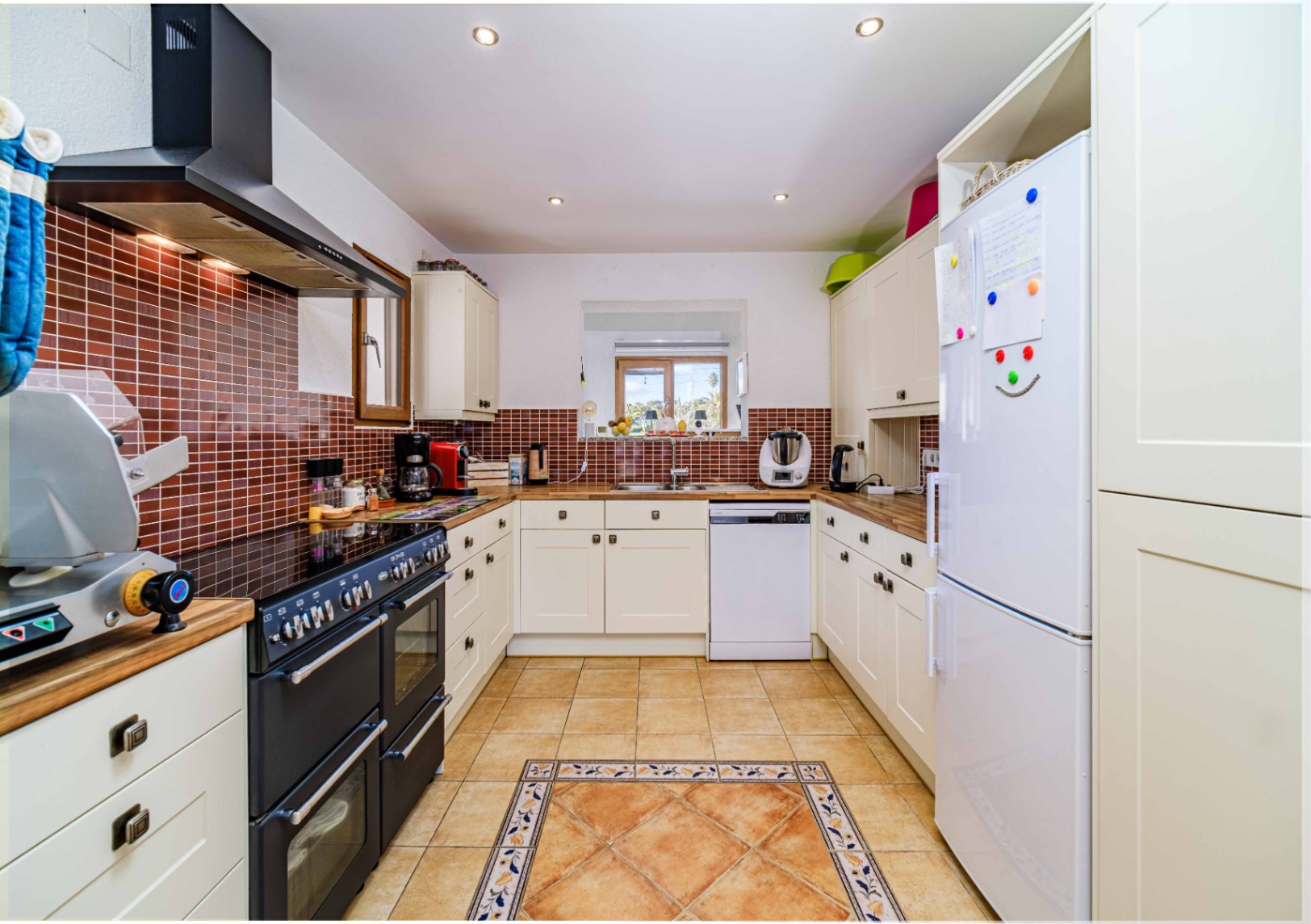 Cocina en forma de U con armarios color crema, encimeras de madera y salpicadero de azulejos marrones. Incluye cocina profesional negra y suelo cerámico con cenefa decorativa.
