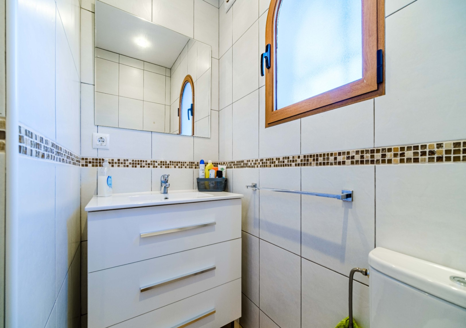 Cuarto de baño con mueble lavabo blanco de tres cajones, espejo rectangular y ventana de arco con marco de madera. Paredes alicatadas en blanco con cenefa de mosaico decorativa.