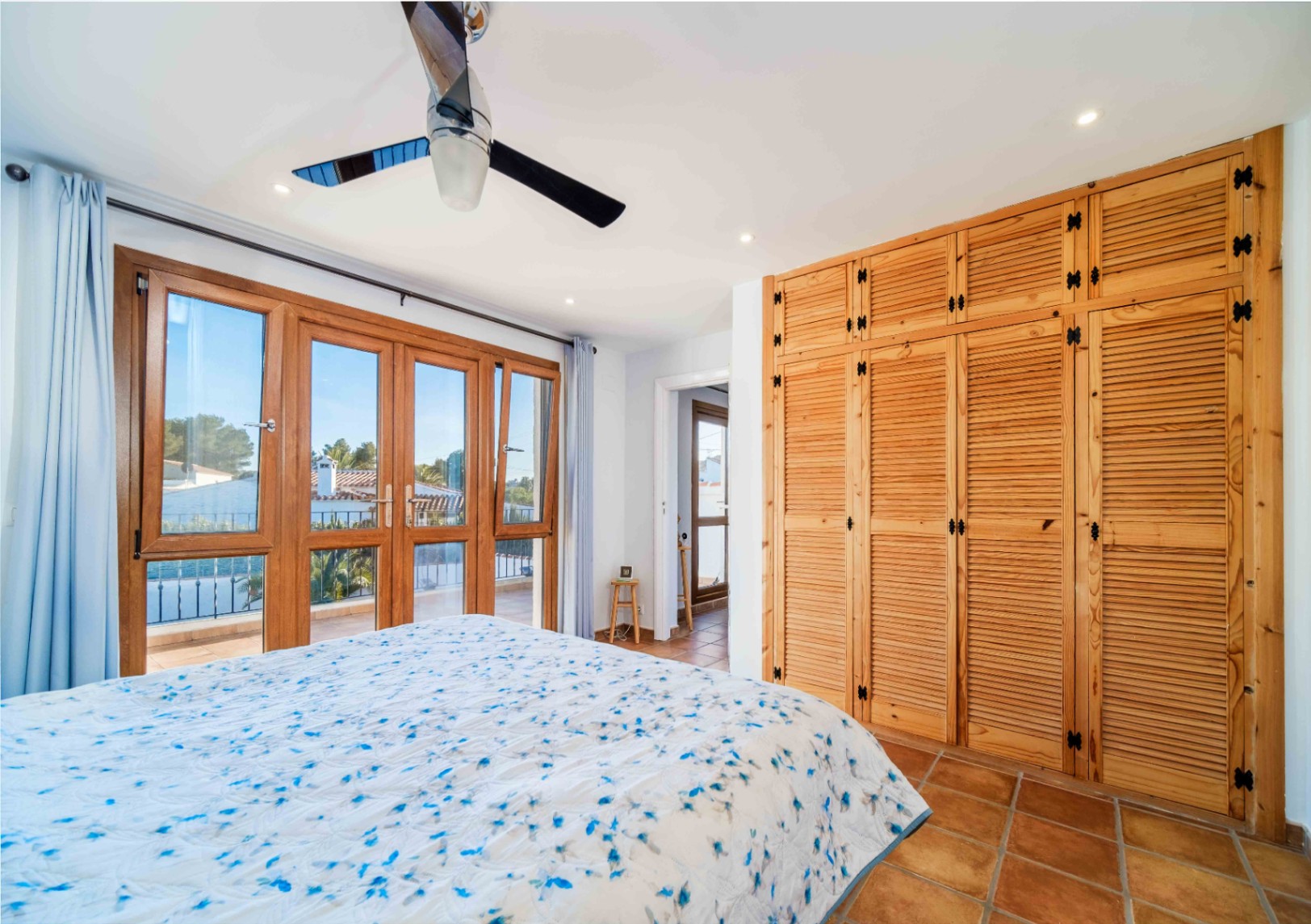 Dormitorio con armarios empotrados de madera tipo persiana, suelo de terracota y acceso directo a la terraza mediante amplios ventanales de madera.