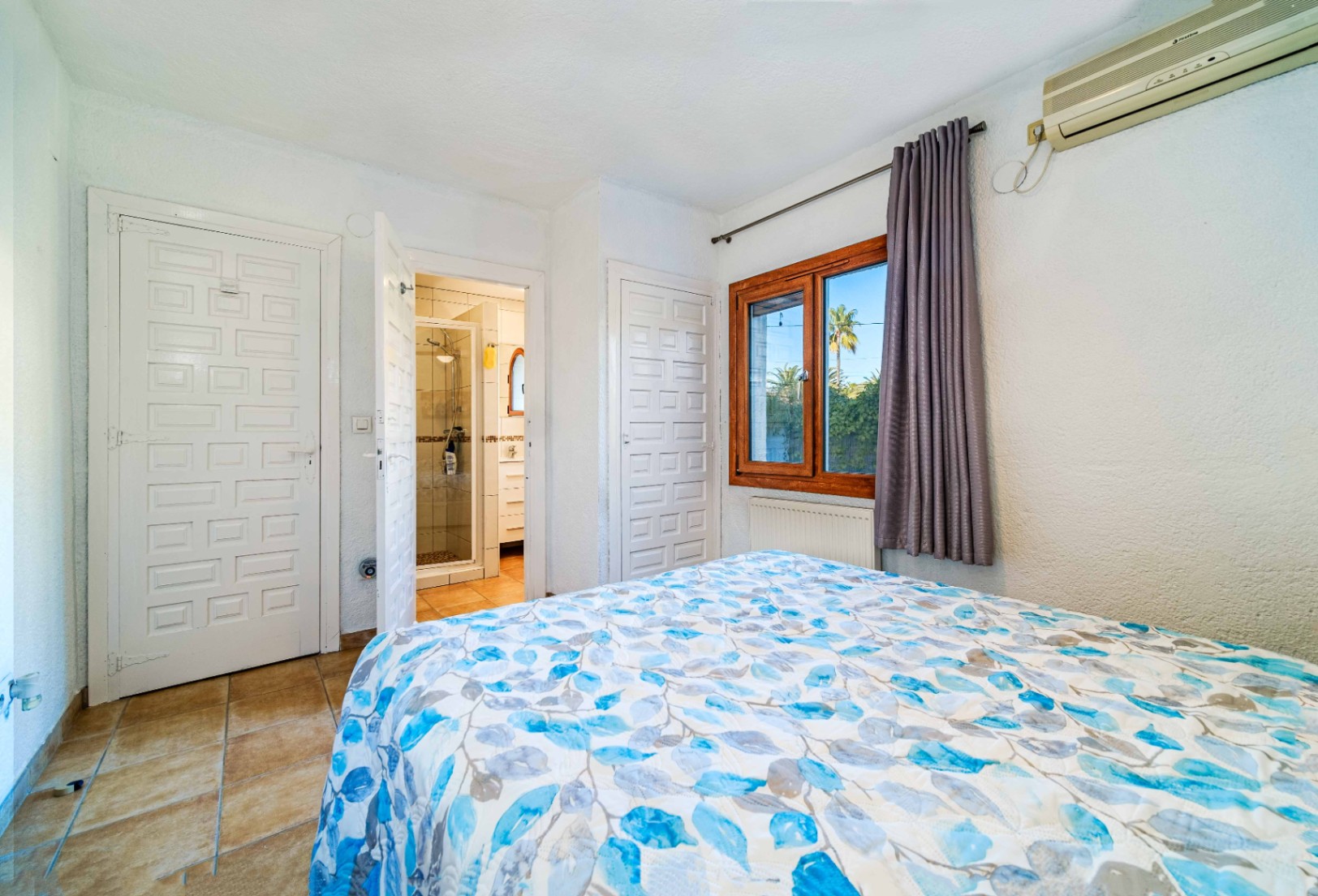 Dormitorio con suelos de terracota, baño en suite con ducha, aire acondicionado y ventana de madera con vistas al jardín.
