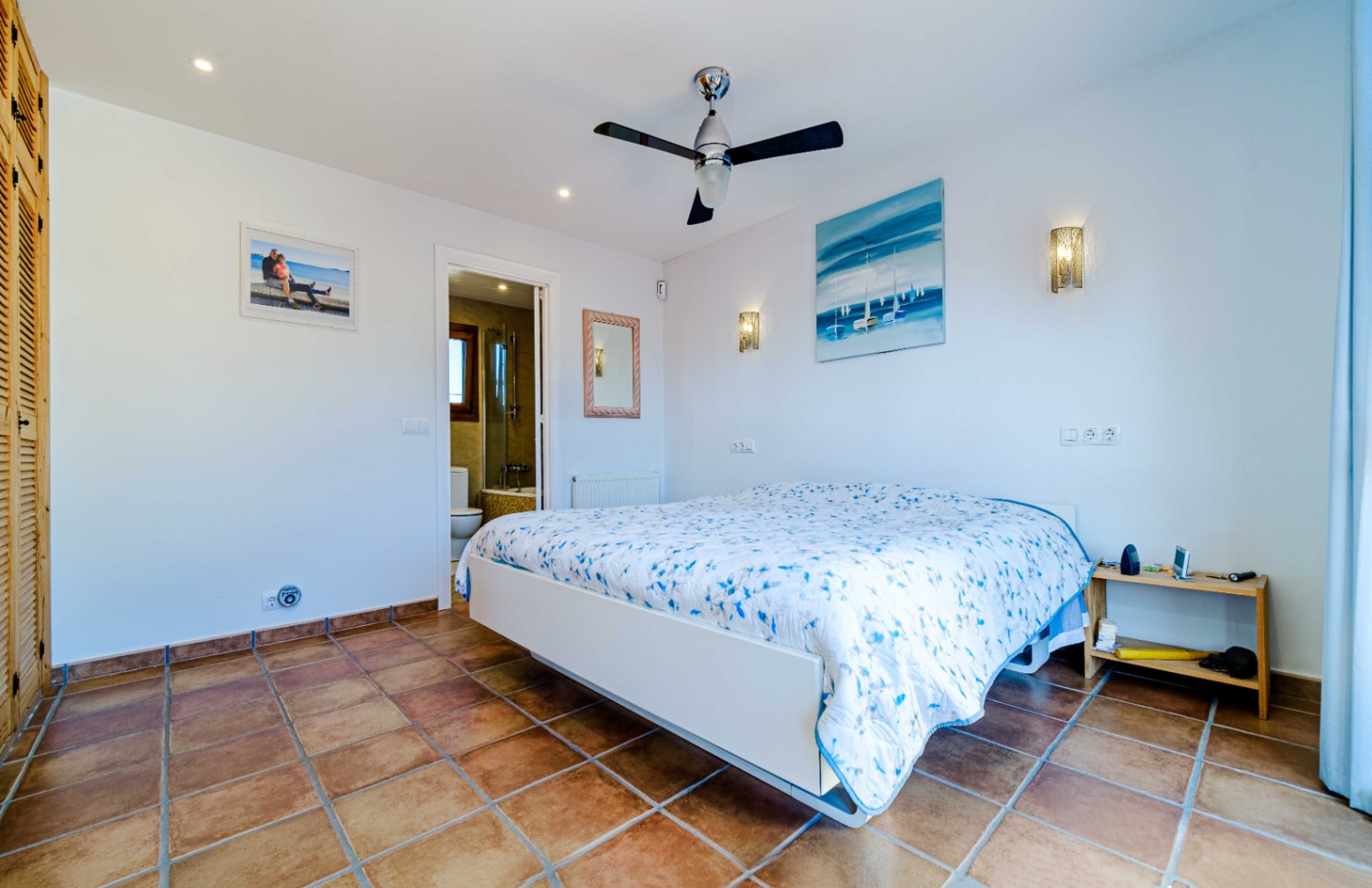 Dormitorio luminoso con suelos de gres rústico, ventilador de techo y baño en suite. Incluye armarios empotrados de madera y apliques de pared decorativos.