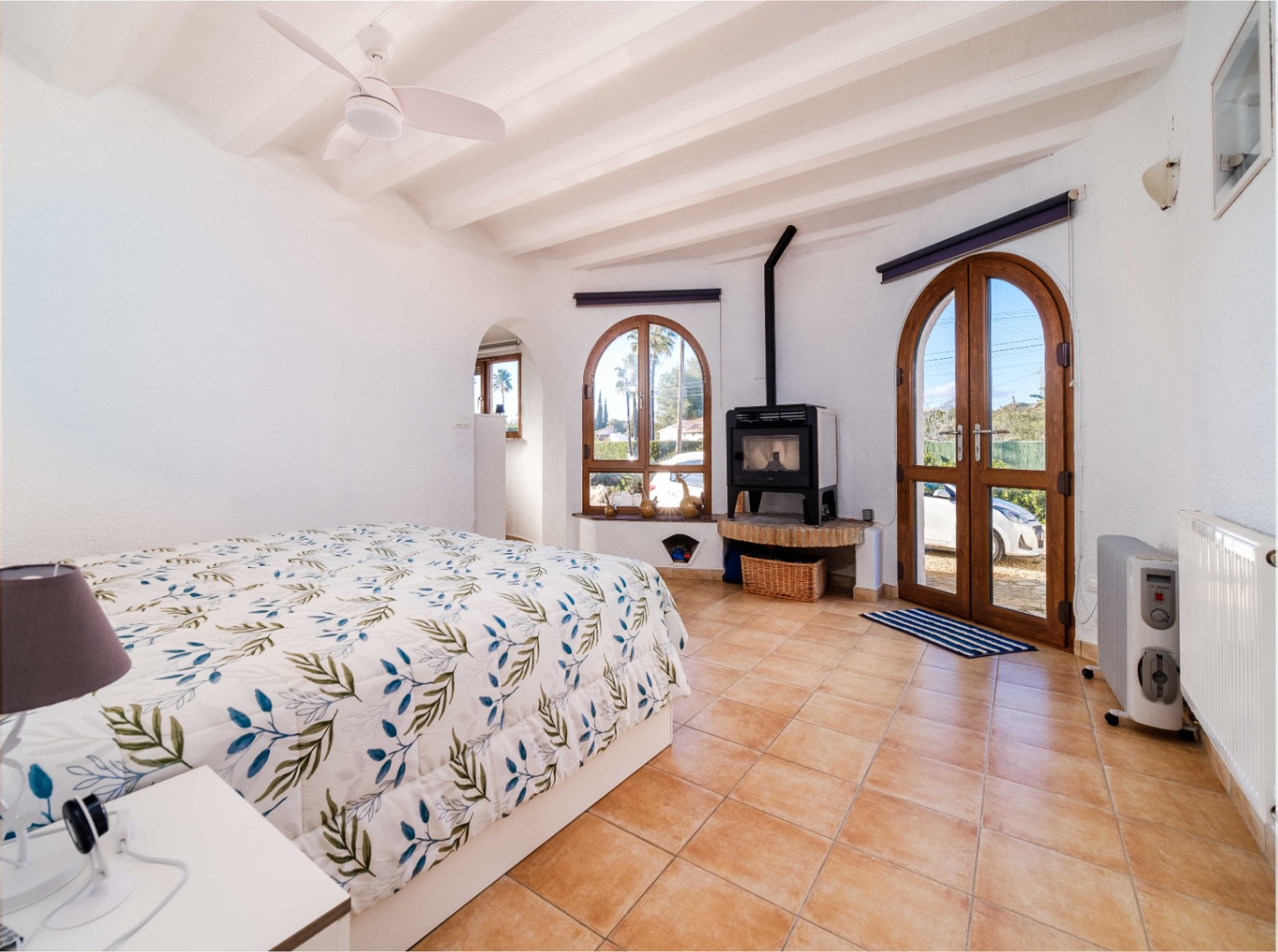 Dormitorio luminoso con techos de vigas blancas, suelo de terracota y una estufa de leña. Incluye acceso exterior mediante puertas de madera en arco y un ventilador de techo.