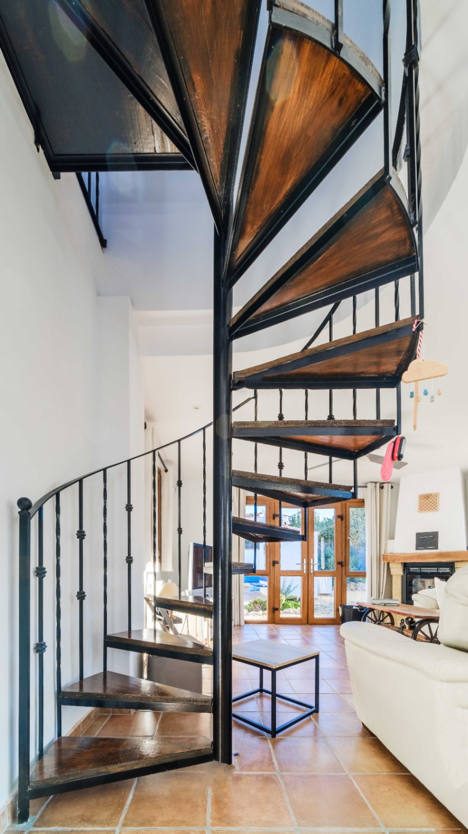 Escalera de caracol con estructura de hierro negro y peldaños de madera oscura, integrada en un salón con suelos de terracota y chimenea al fondo.