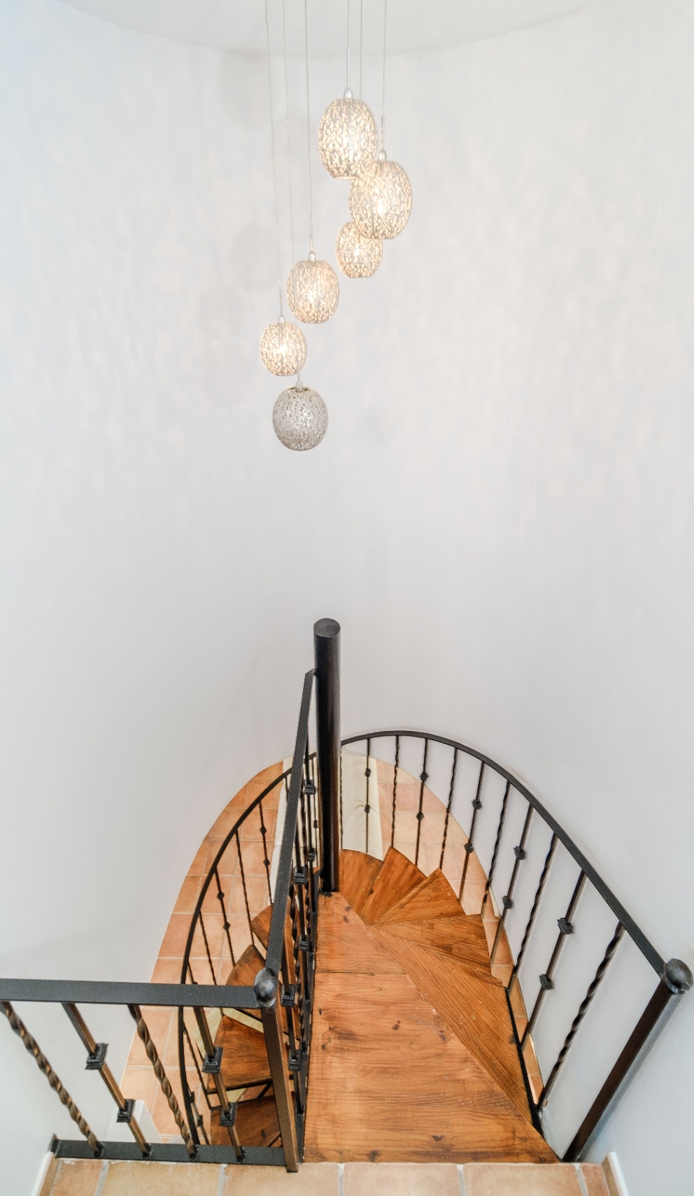 Escalera de caracol con peldaños de madera maciza y barandilla de hierro forjado negro, iluminada por un conjunto de lámparas colgantes esféricas de diseño.
