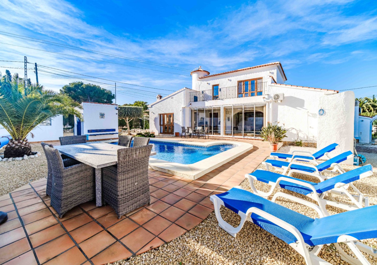 Exterior de la villa con piscina, zona de comedor al aire libre y hamacas, destacando la fachada blanca tradicional y la naya acristalada bajo el sol de Jávea.