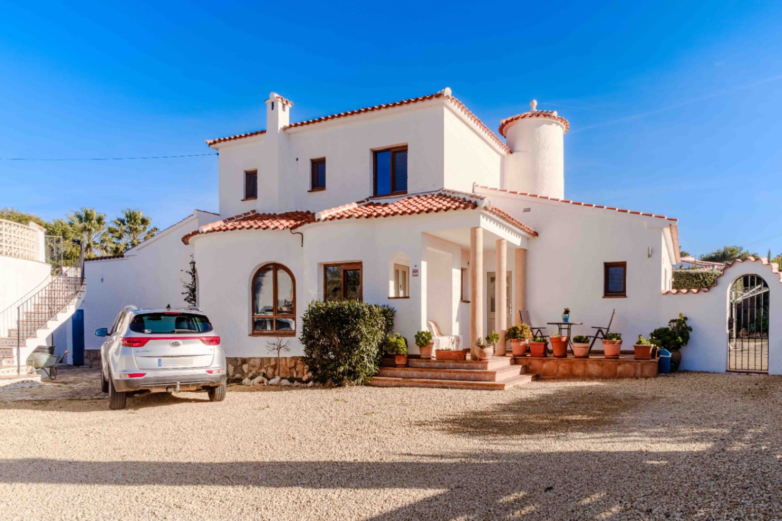 Fachada principal de la villa con arquitectura mediterránea, muros blancos, torre circular y porche con columnas sobre una amplia zona de grava.