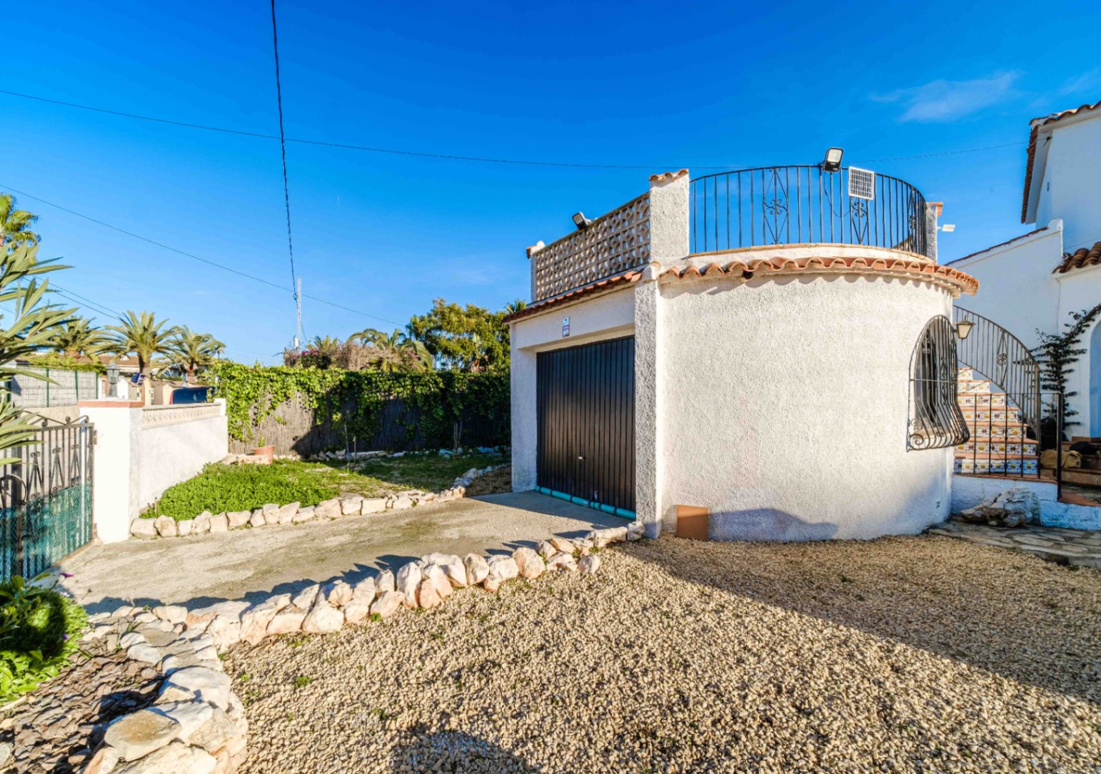 Garaje independiente con fachada curva de estuco blanco, puerta metálica oscura y terraza superior con barandilla de hierro forjado y panel solar.