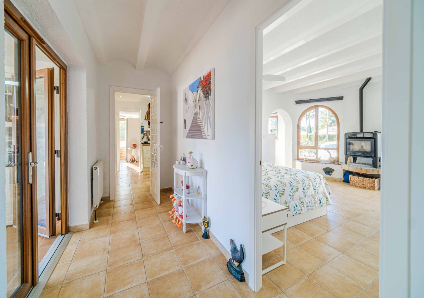 Pasillo con suelos cerámicos y techos de vigas blancas que conecta con un dormitorio luminoso equipado con chimenea de leña y ventanal de arco.