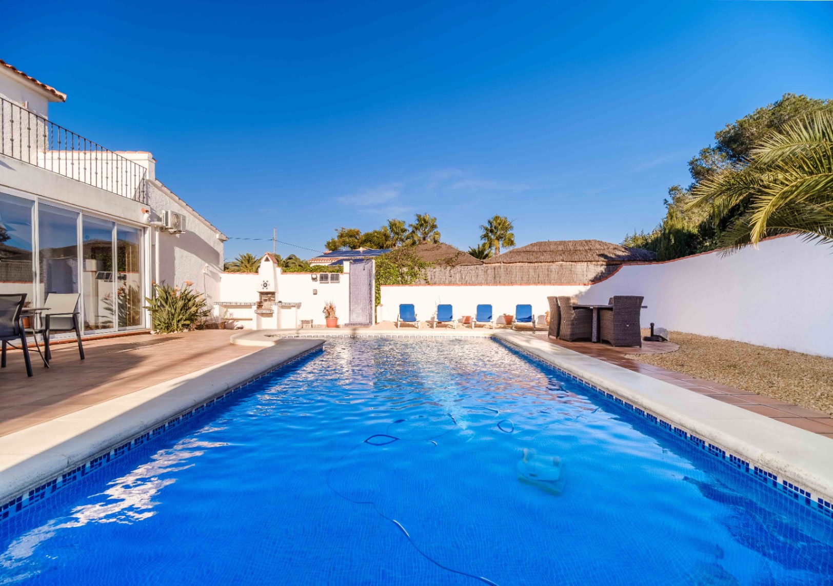Piscina rectangular de gresite azul rodeada de una terraza de terracota, con zona de barbacoa integrada, comedor exterior y paneles solares visibles al fondo.