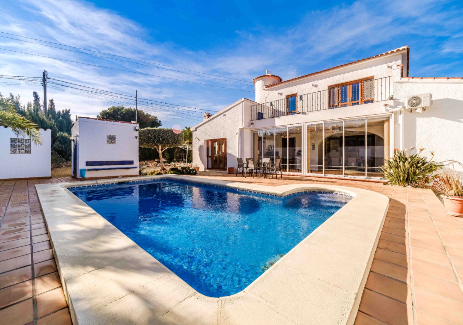 Piscina rodeada de una amplia terraza de terracota, con vistas a la fachada blanca de la villa y su naya acristalada frente al jardín mediterráneo.