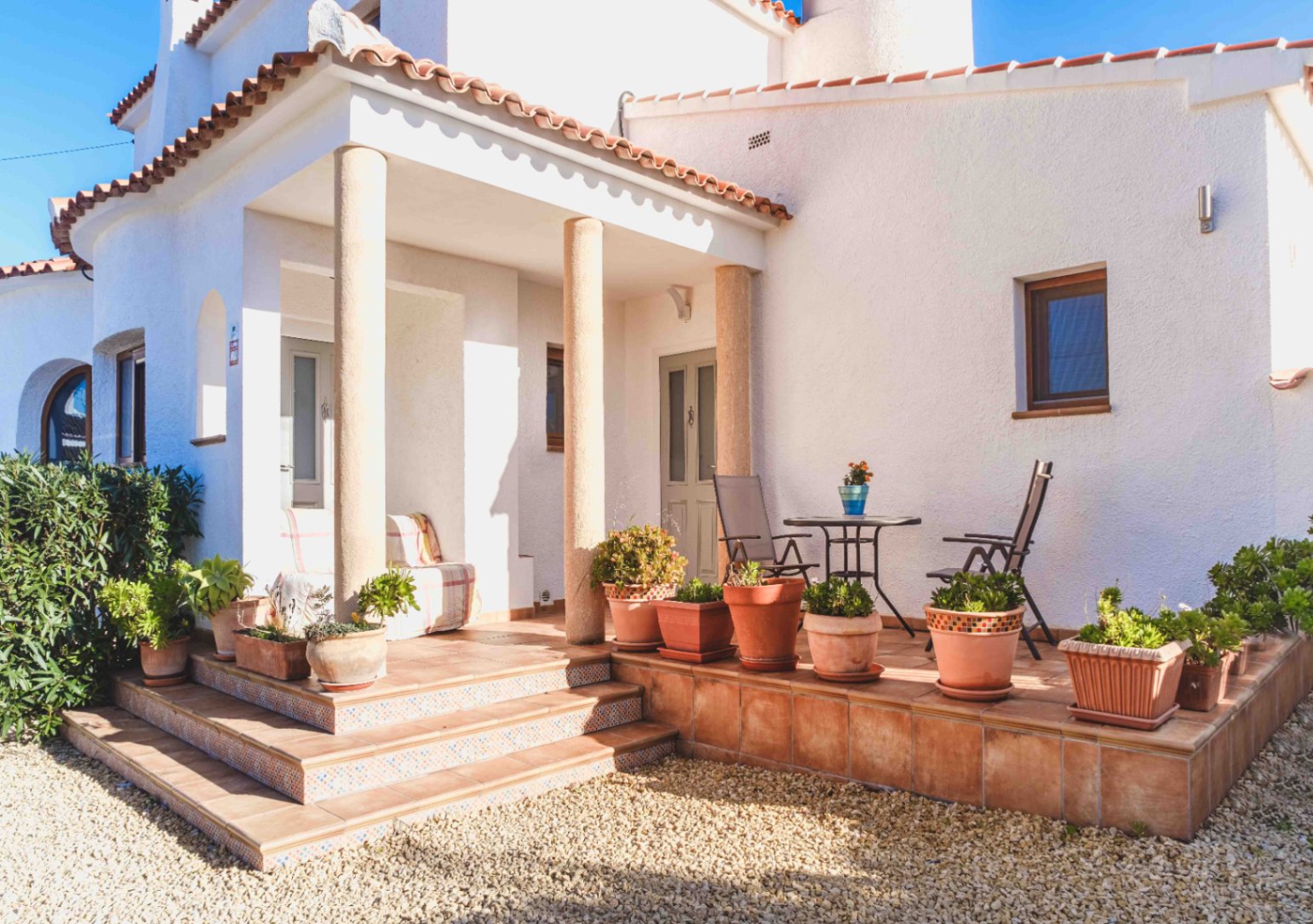 Porche de entrada con columnas de piedra, suelos de terracota y escalones decorados. La fachada blanca se complementa con macetas de barro y una zona de estar exterior.