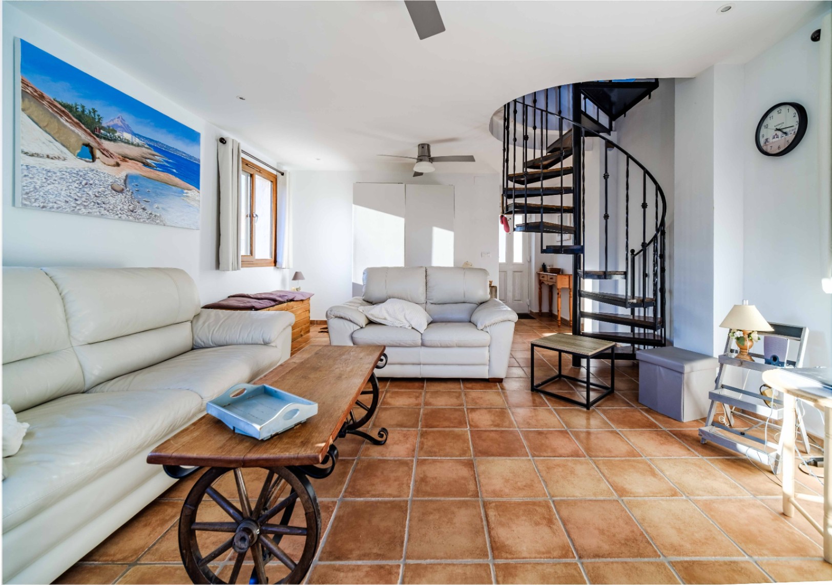 Salón con suelos de terracota, sofás de cuero blanco y una singular mesa de centro con ruedas de madera. Destaca la escalera de caracol metálica y la abundante luz natural.