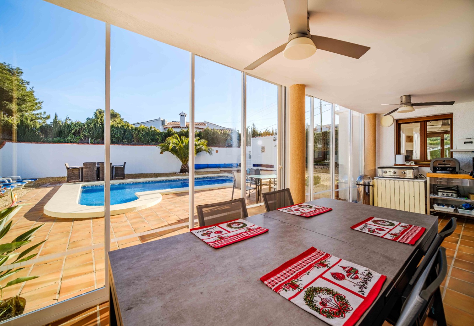 Terraza acristalada con amplio comedor, ventiladores de techo y vistas panorámicas a la piscina privada y zona de cocina exterior.