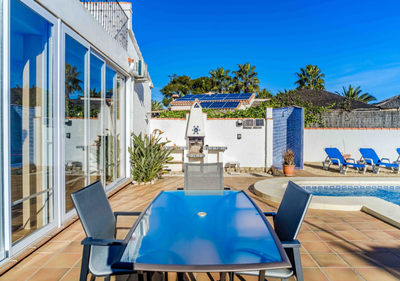 Terraza exterior con comedor de cristal, barbacoa de obra y ducha de mosaico azul junto a la piscina. Destacan los paneles solares y los amplios ventanales.