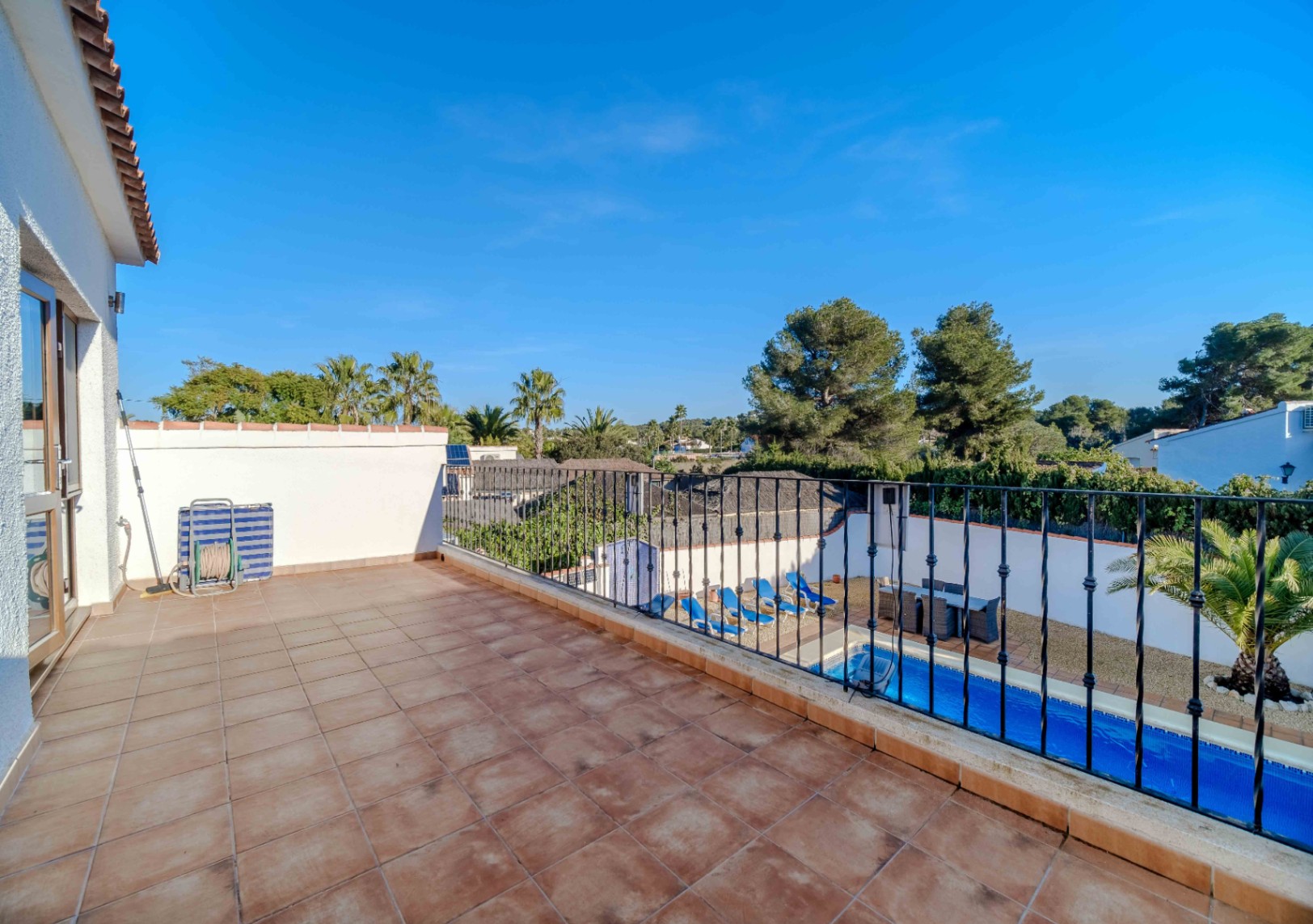 Terraza superior con suelo de gres cerámico y barandilla de forja, con vistas despejadas a la piscina de agua salada y al entorno arbolado de Jávea.