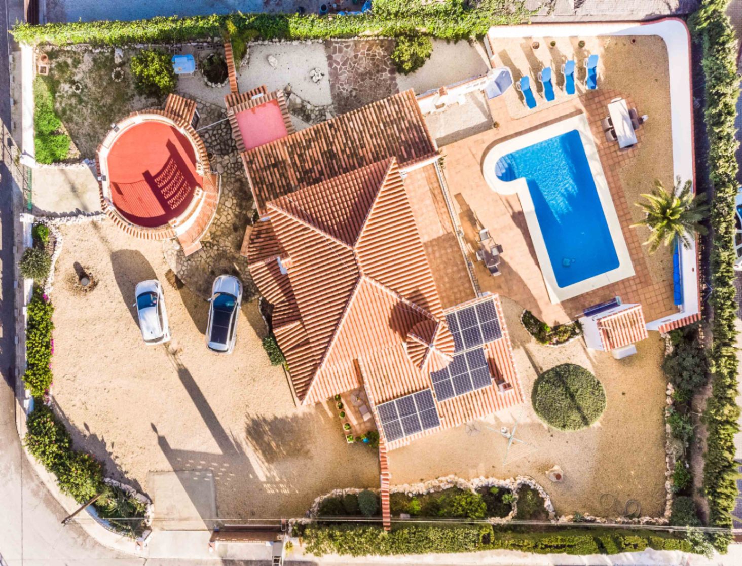 Vista aérea de la villa que muestra los paneles solares en el tejado, la piscina privada con solárium y la distribución de la parcela con zona de aparcamiento.