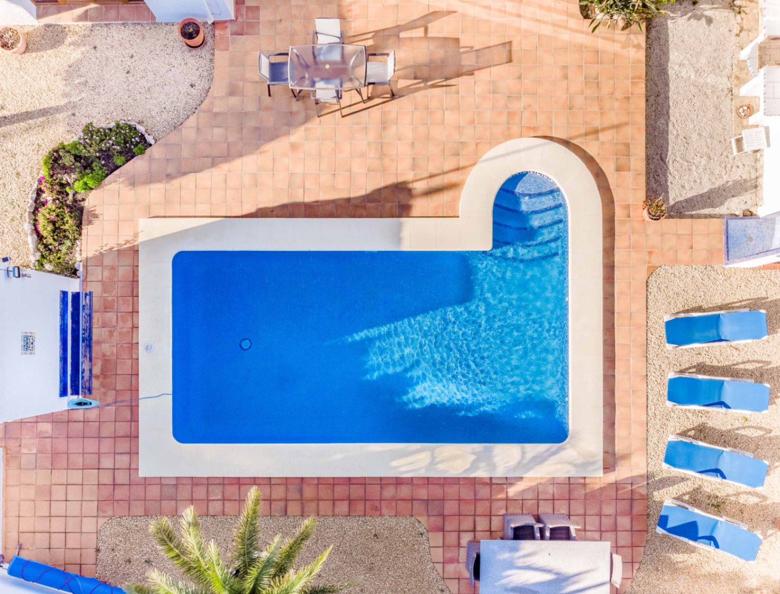 Vista cenital de la piscina rectangular con escalera romana, rodeada de una amplia terraza de baldosas de terracota y zonas de descanso con tumbonas azules.