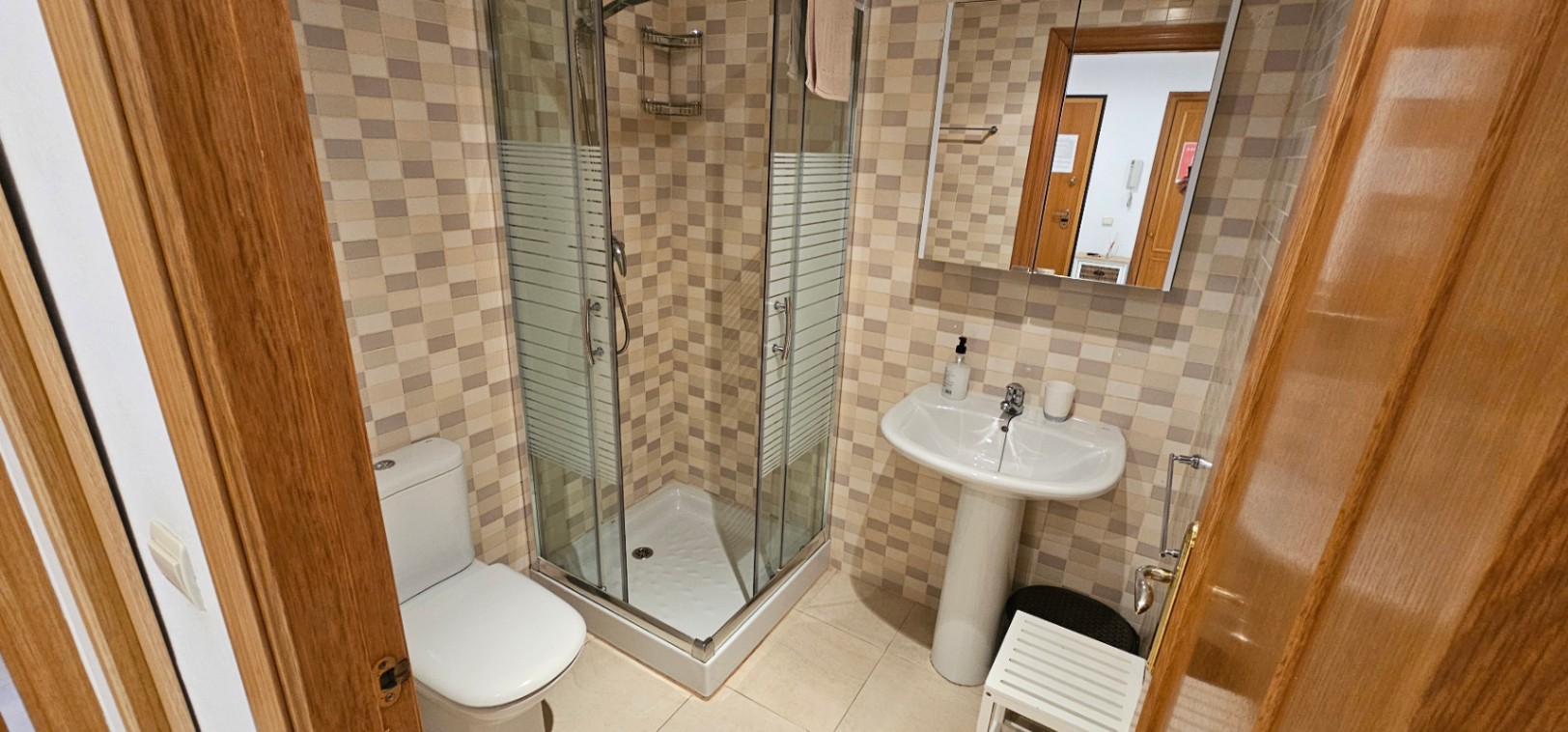 Baño funcional con ducha de esquina acristalada, lavabo de pedestal y paredes revestidas con azulejos de mosaico en tonos tierra.