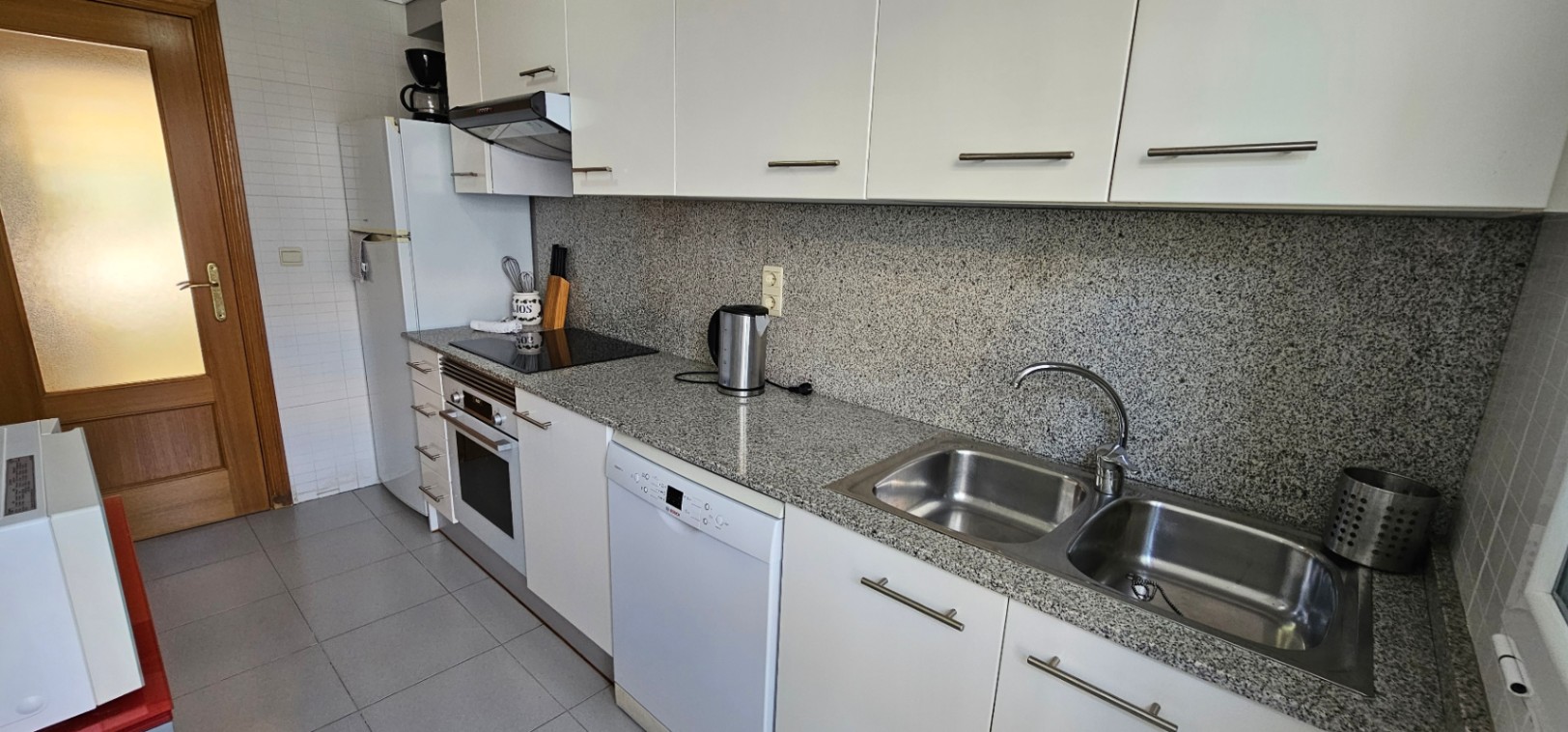 Cocina equipada con armarios blancos, encimera de granito gris, electrodomésticos integrados y fregadero doble de acero inoxidable.