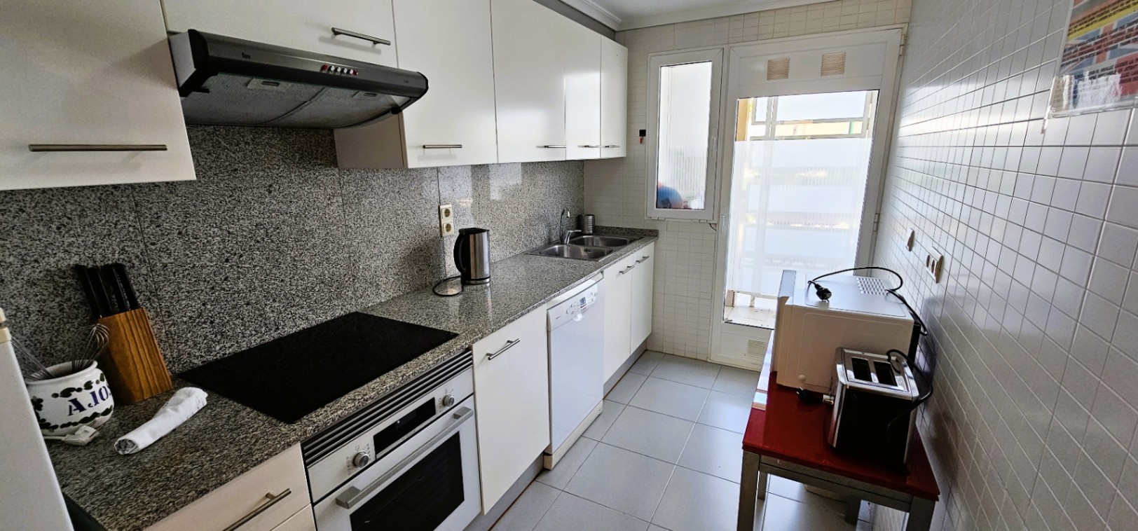 Cocina independiente equipada con encimera de granito, muebles blancos, horno, placa vitrocerámica y acceso directo al exterior a través de una puerta acristalada.
