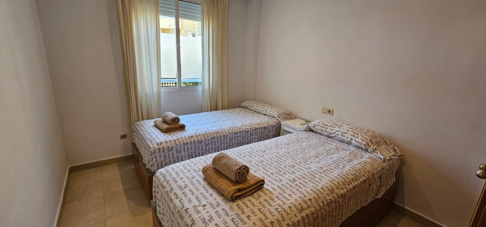 Dormitorio luminoso con dos camas individuales, suelos de gres y ventana exterior con cortinas claras que aporta luz natural.