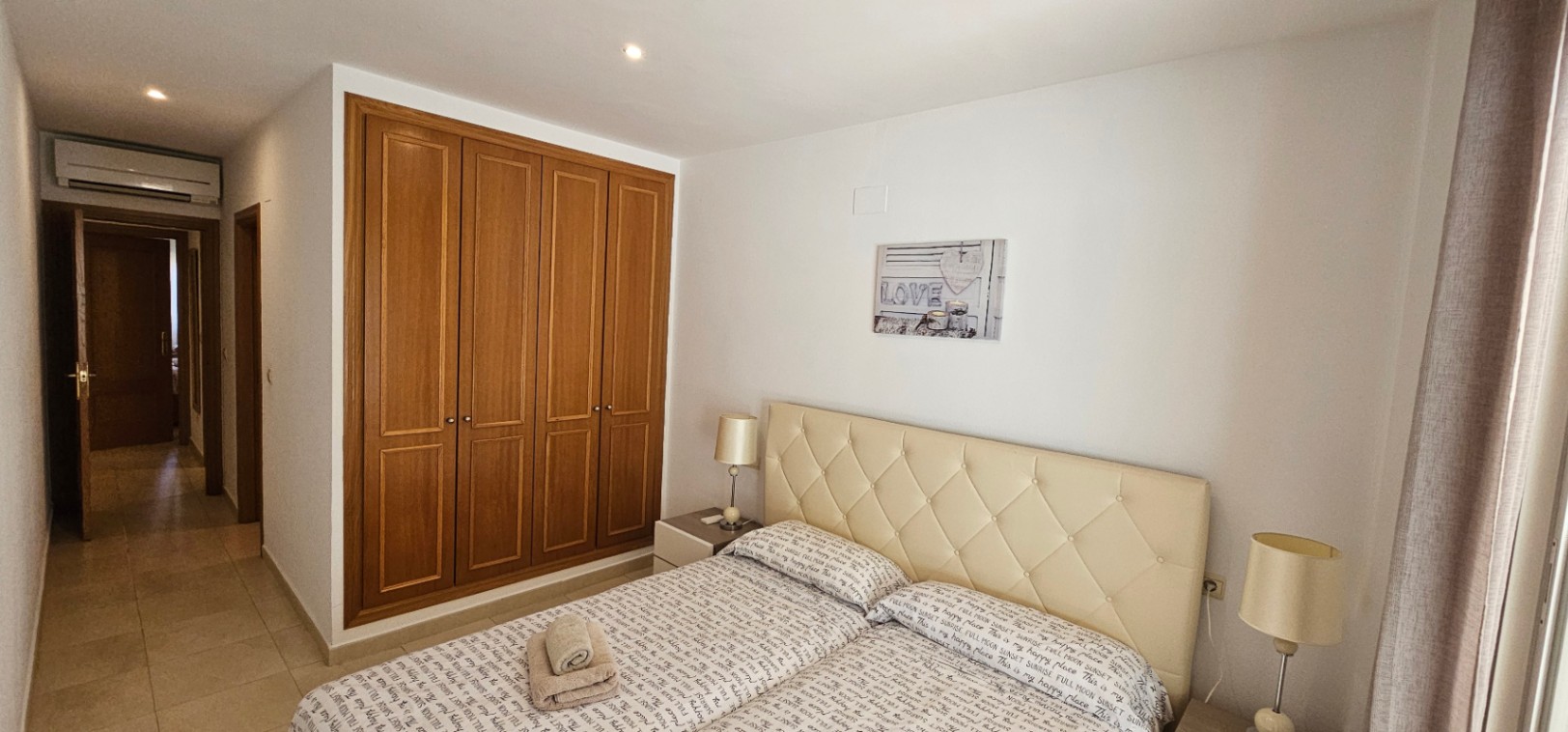 Dormitorio principal con armarios empotrados de madera, cabecero acolchado en tono crema y aire acondicionado integrado sobre la puerta de acceso.