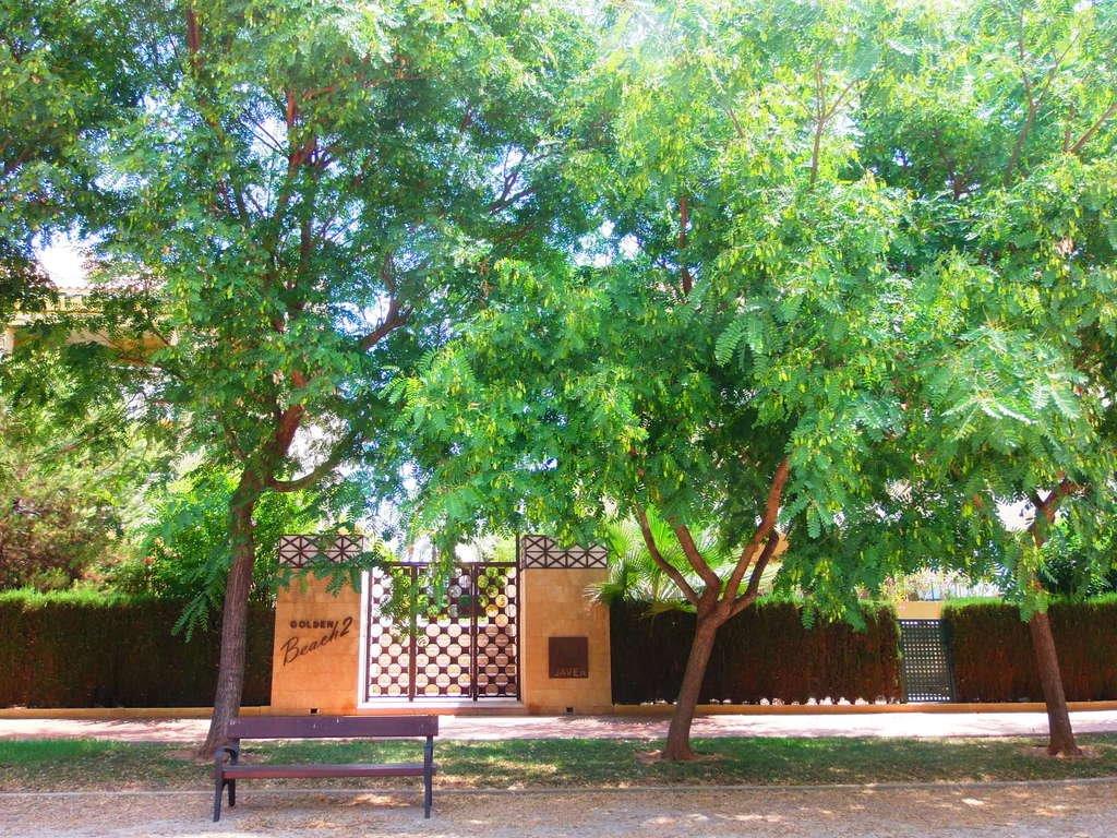 Entrada principal a la urbanización Golden Beach 2, con puertas de hierro forjado, pilares de piedra y frondosos árboles que ofrecen sombra sobre un banco de descanso.