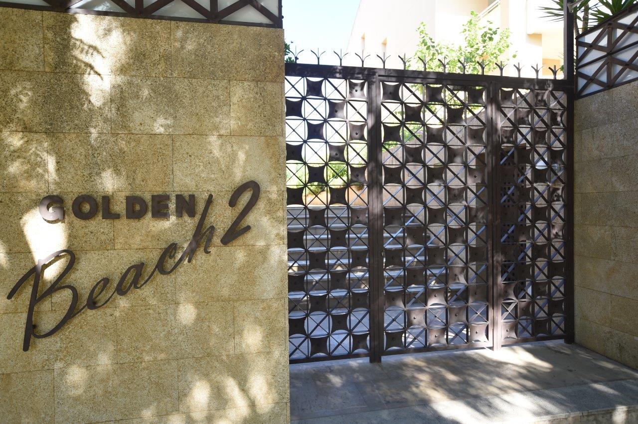 Entrada principal de la urbanización Golden Beach 2, con muro de piedra natural, rotulación metálica y puerta de seguridad con diseño geométrico.