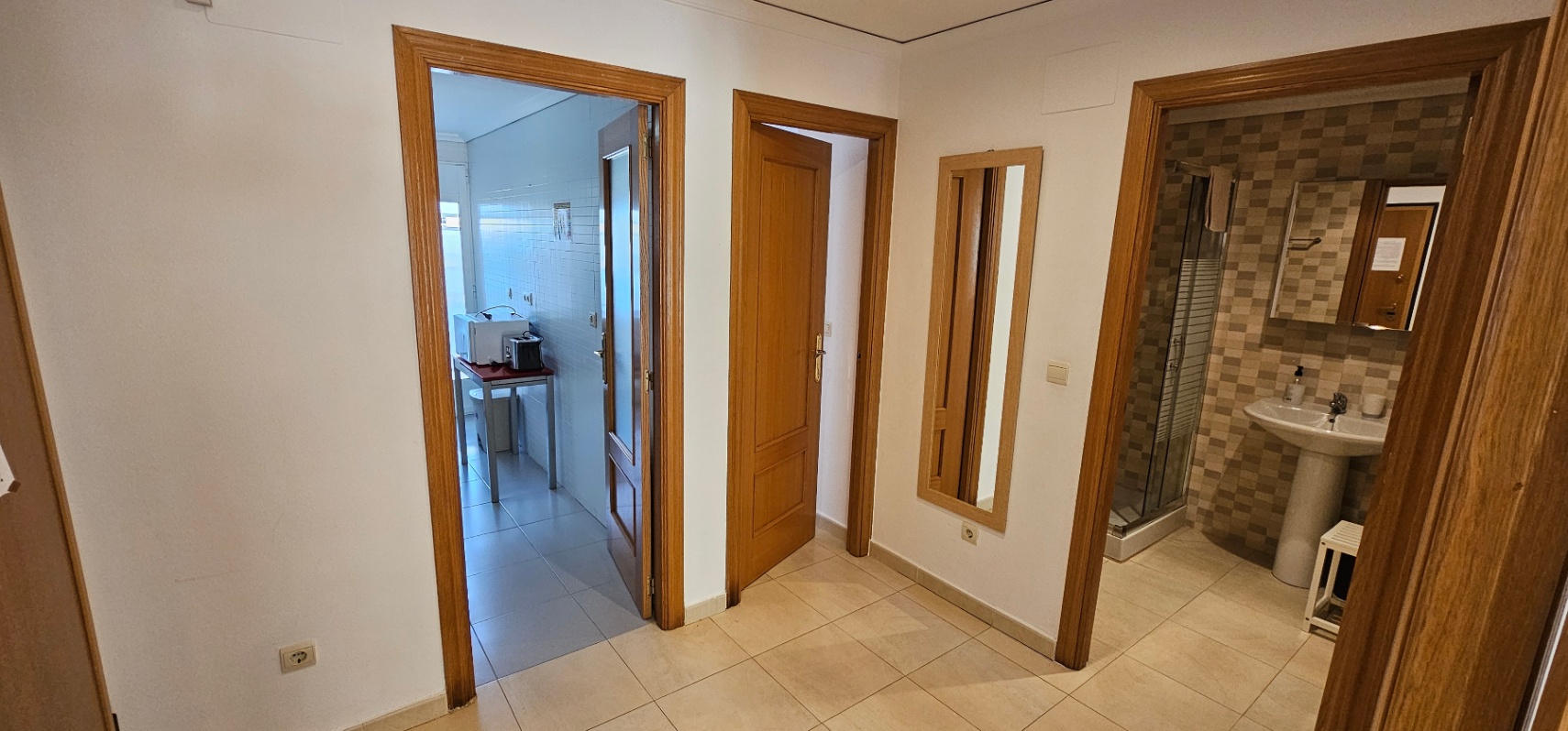 Pasillo distribuidor con marcos de madera que conecta la cocina, un dormitorio y un baño con ducha de azulejos tipo mosaico.