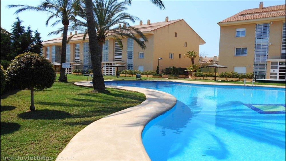 Piscina comunitaria de forma orgánica rodeada de amplias zonas de césped, palmeras maduras y edificios residenciales de estilo mediterráneo en Jávea.