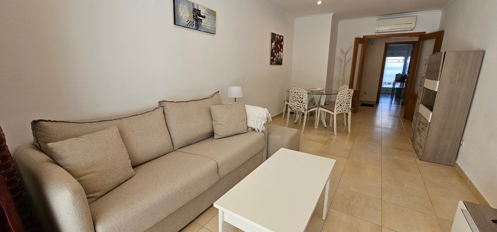 Salón comedor luminoso con sofá beige, mesa de centro blanca y zona de comedor con sillas de diseño. Incluye aire acondicionado y suelos cerámicos claros.