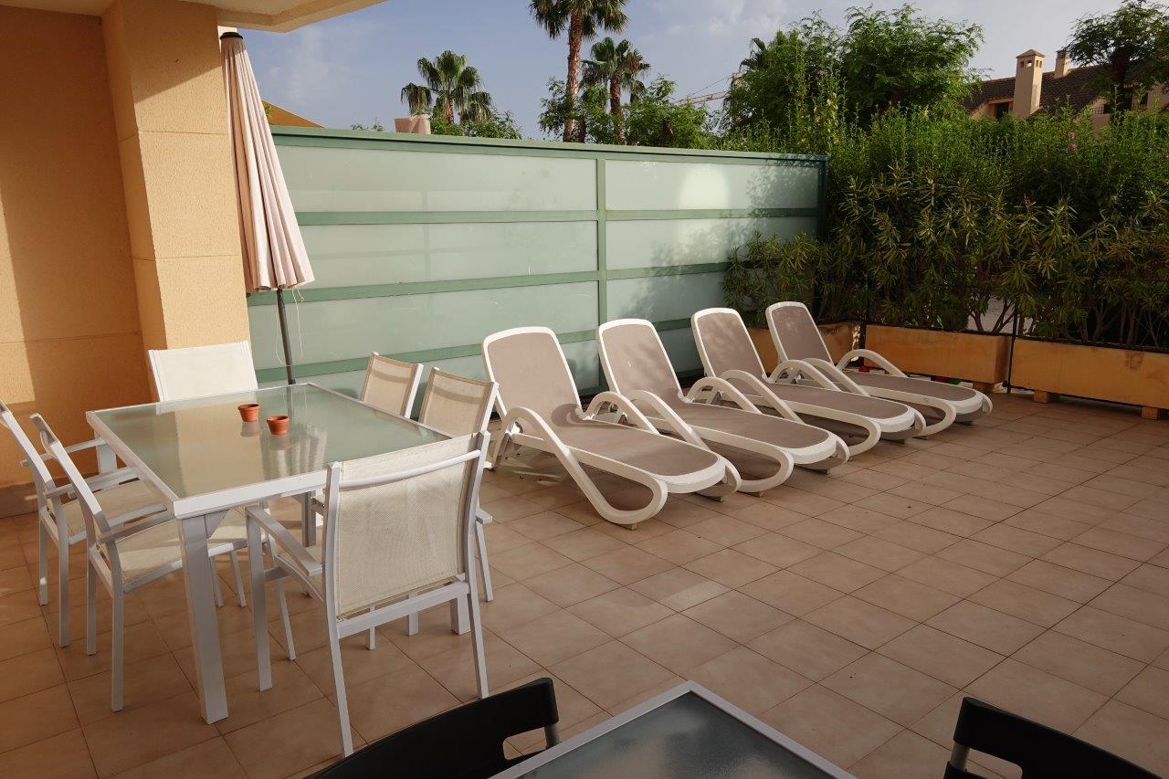 Terraza privada con suelo cerámico, equipada con zona de comedor exterior y tumbonas, protegida por un panel de cristal translúcido y vegetación perimetral.