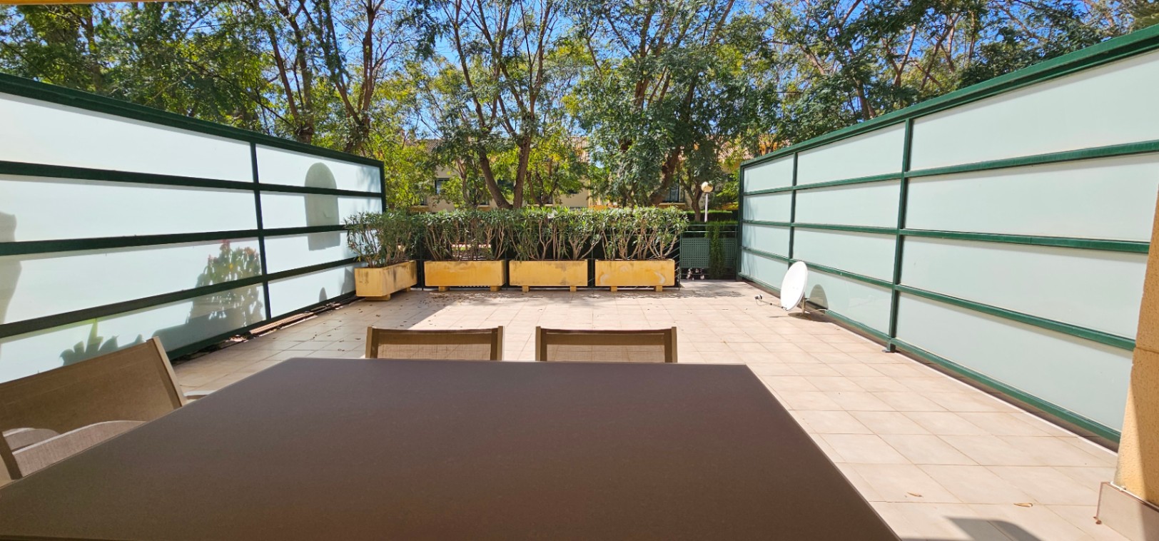 Terraza privada con suelo cerámico, paneles laterales de cristal traslúcido para mayor intimidad y jardineras con arbustos bajo la sombra de árboles maduros.